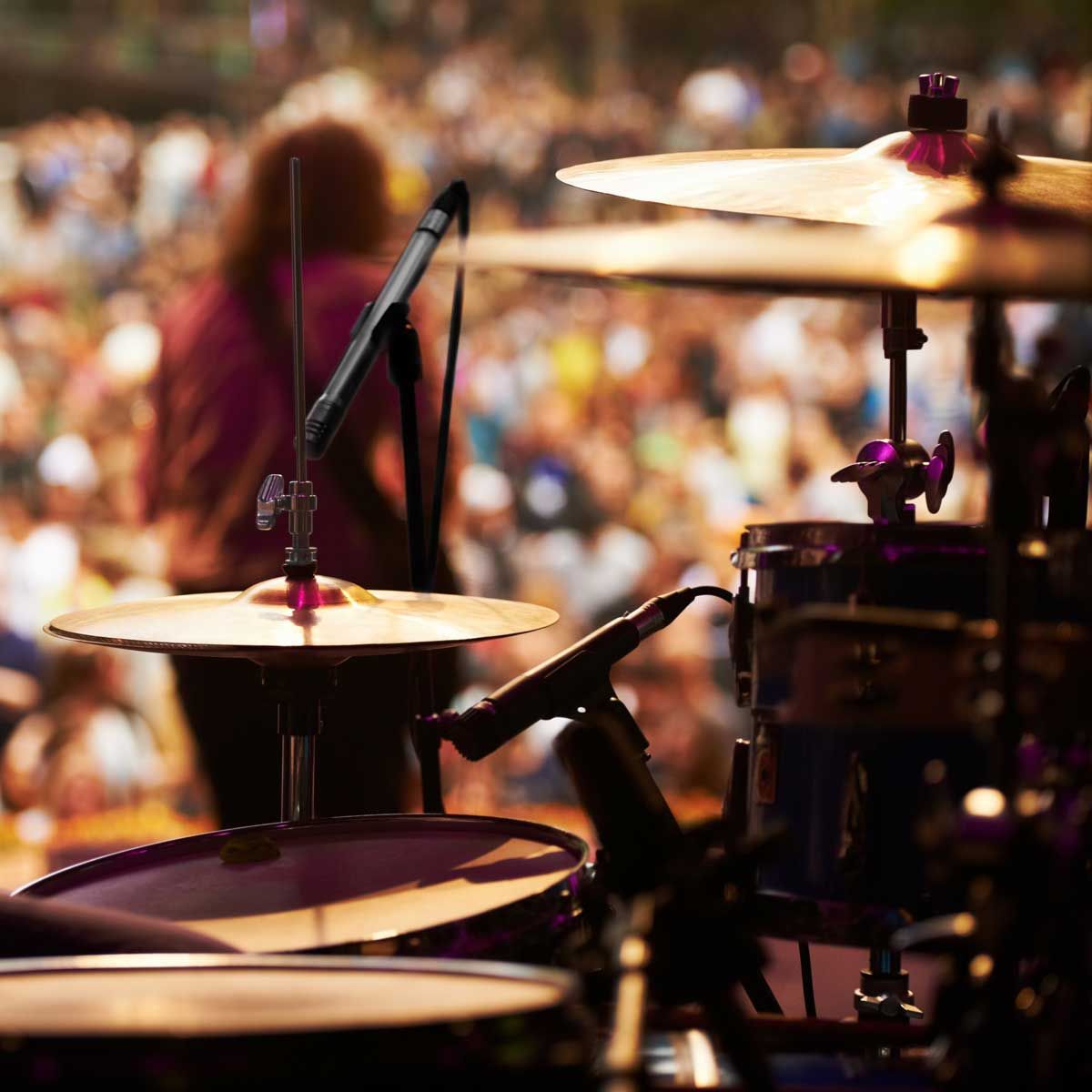 Drum set on stage, mic'd up, with a musician blurred in the background, in front of a crowd.