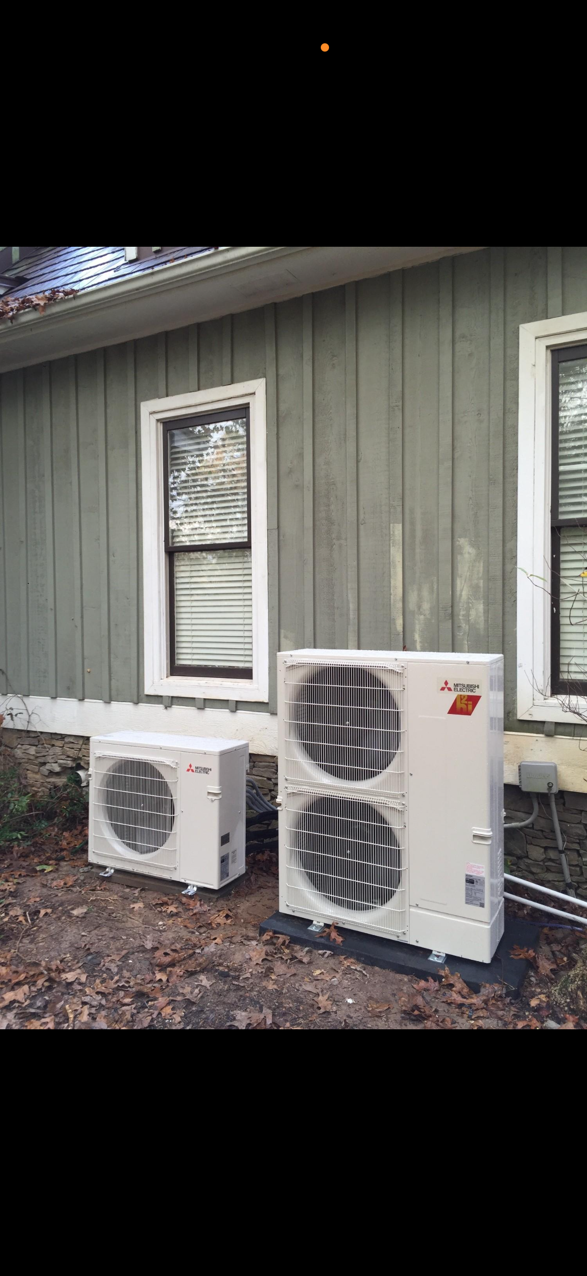 A gray outdoor air conditioning unit sits on a concrete pad against a light-colored brick wall above a patch of grass.