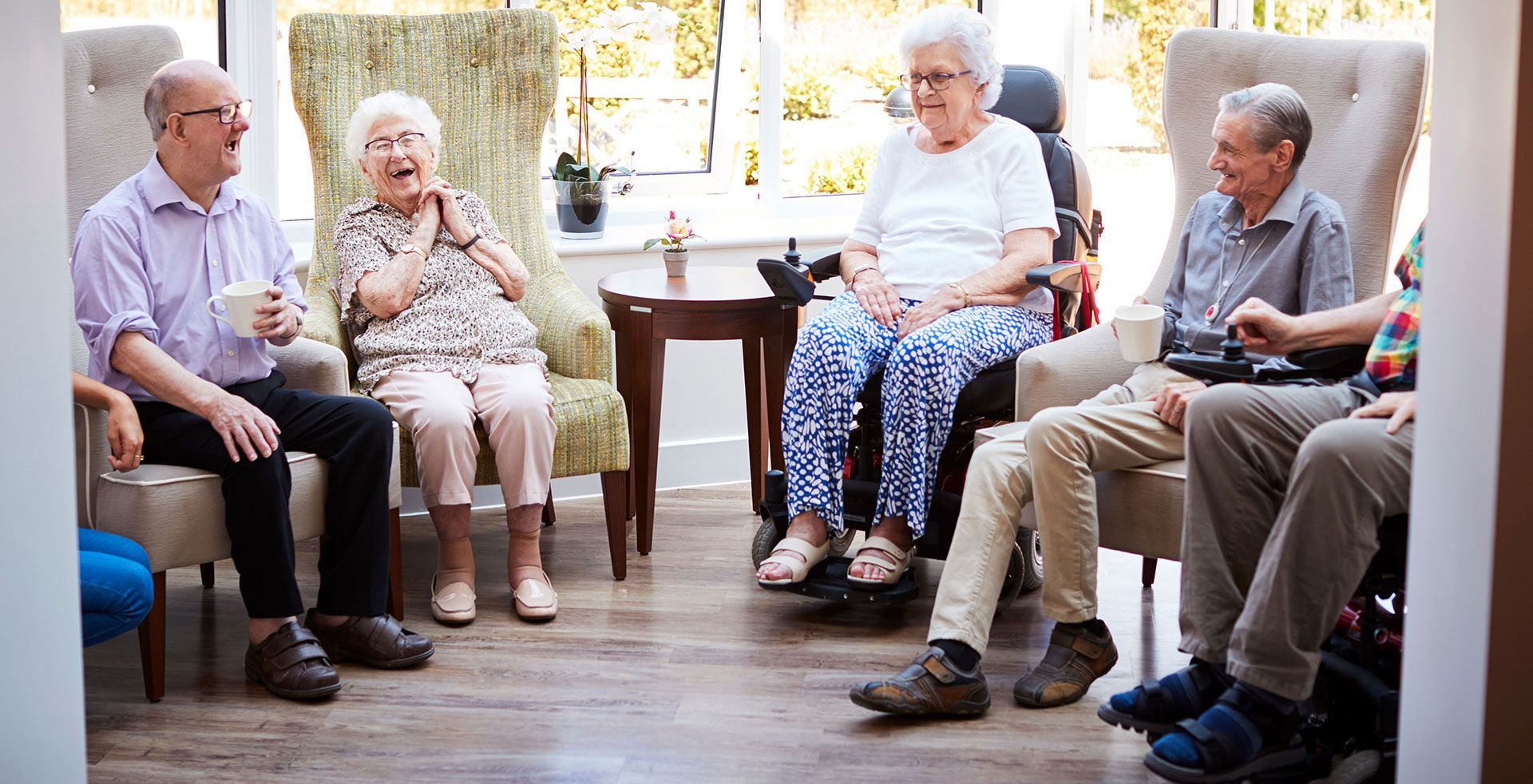 Male And Female Residents Sitting In Chairs And Talking In Lounge Of Retirement Home