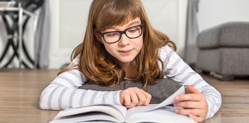 Young girl wearing glasses reading book while lying on floor at home
