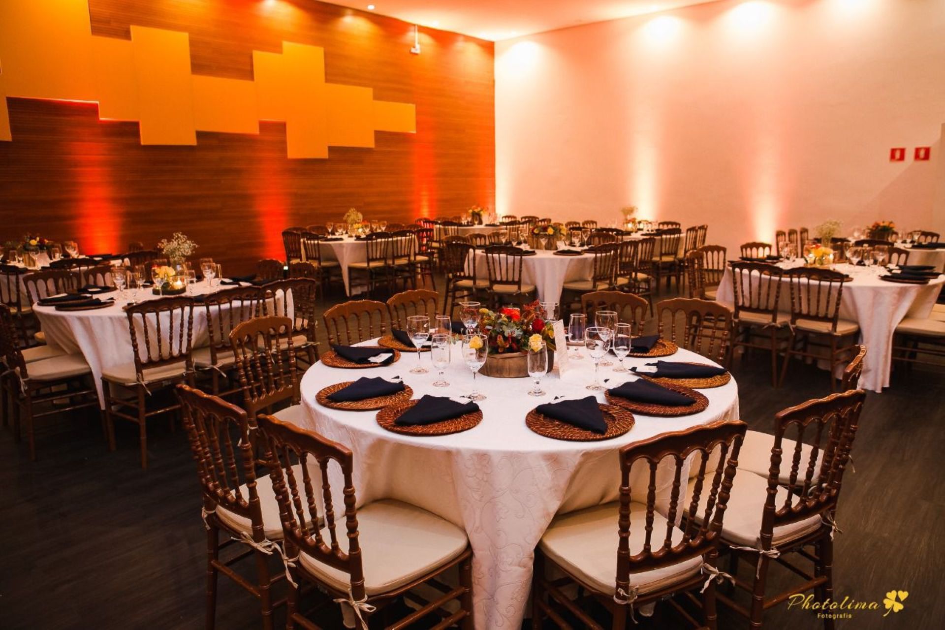 Ambiente formal para evento, com mesas redondas, toalhas de mesa brancas, cadeiras marrons e arranjos de mesa em uma sala com iluminação aconchegante.