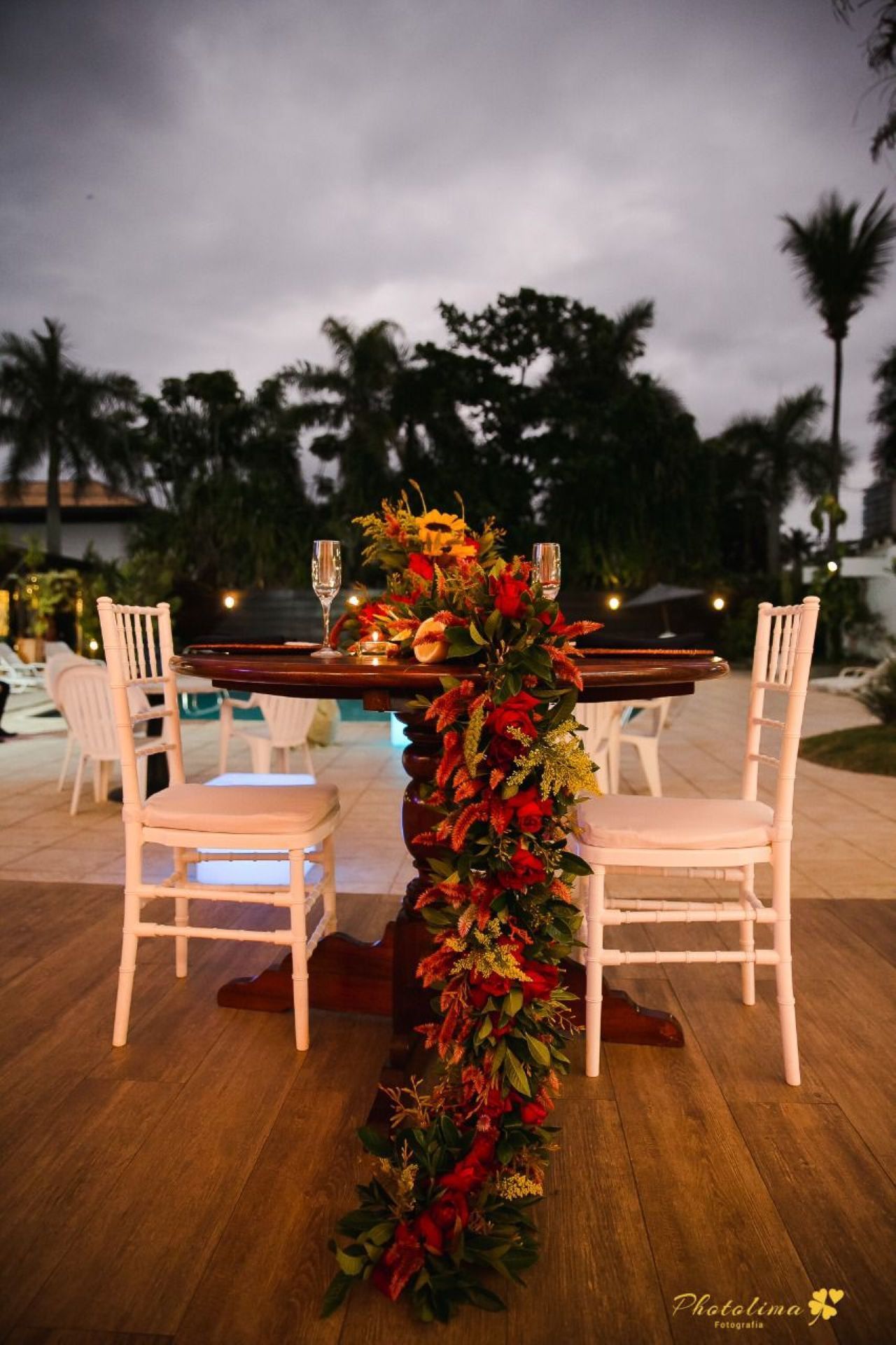 Mesa romântica posta com arranjo floral, duas cadeiras e vista para a piscina.