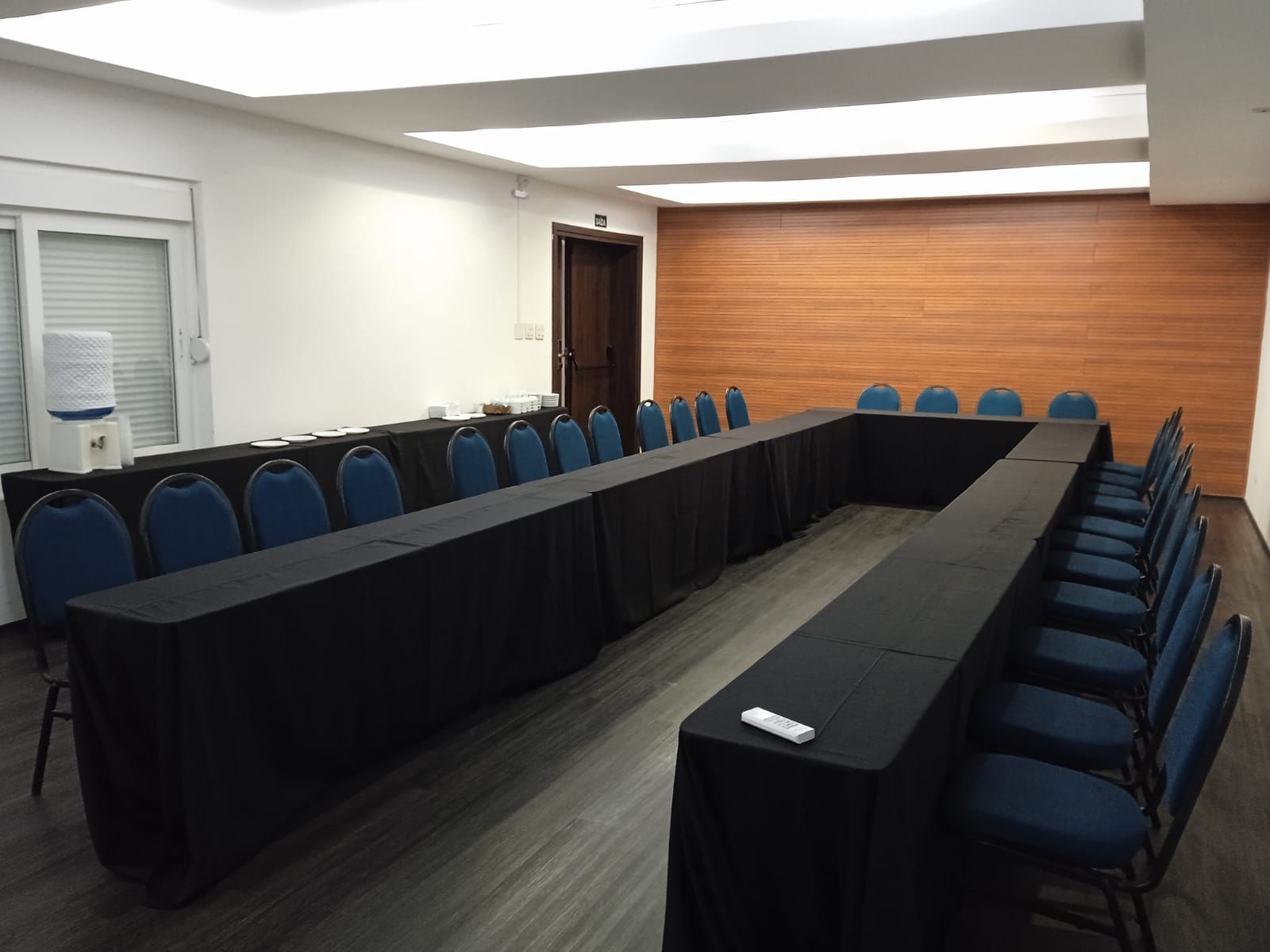 Sala de conferências com mesa em formato de U coberta com toalha preta, cadeiras azuis e parede com painéis de madeira.