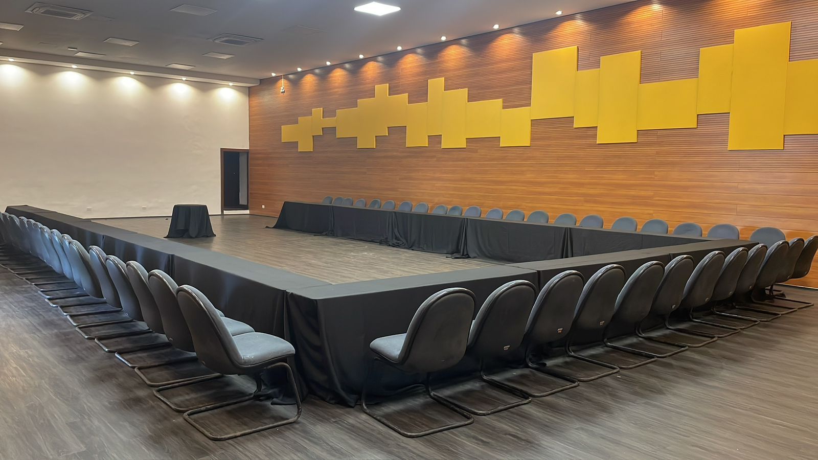 Sala de conferências com mesa em formato de U coberta com toalha preta; cadeiras cinza voltadas para o centro. Paredes amarelas com painéis de madeira.