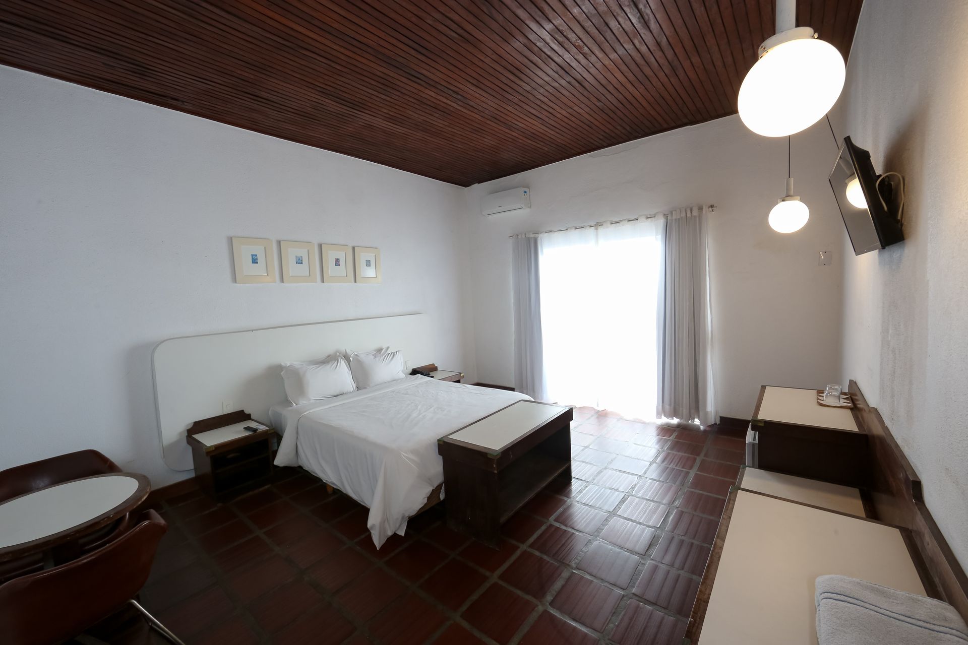 Quarto com paredes brancas, teto de madeira, cama com lençóis brancos, janela grande e piso de azulejo marrom.