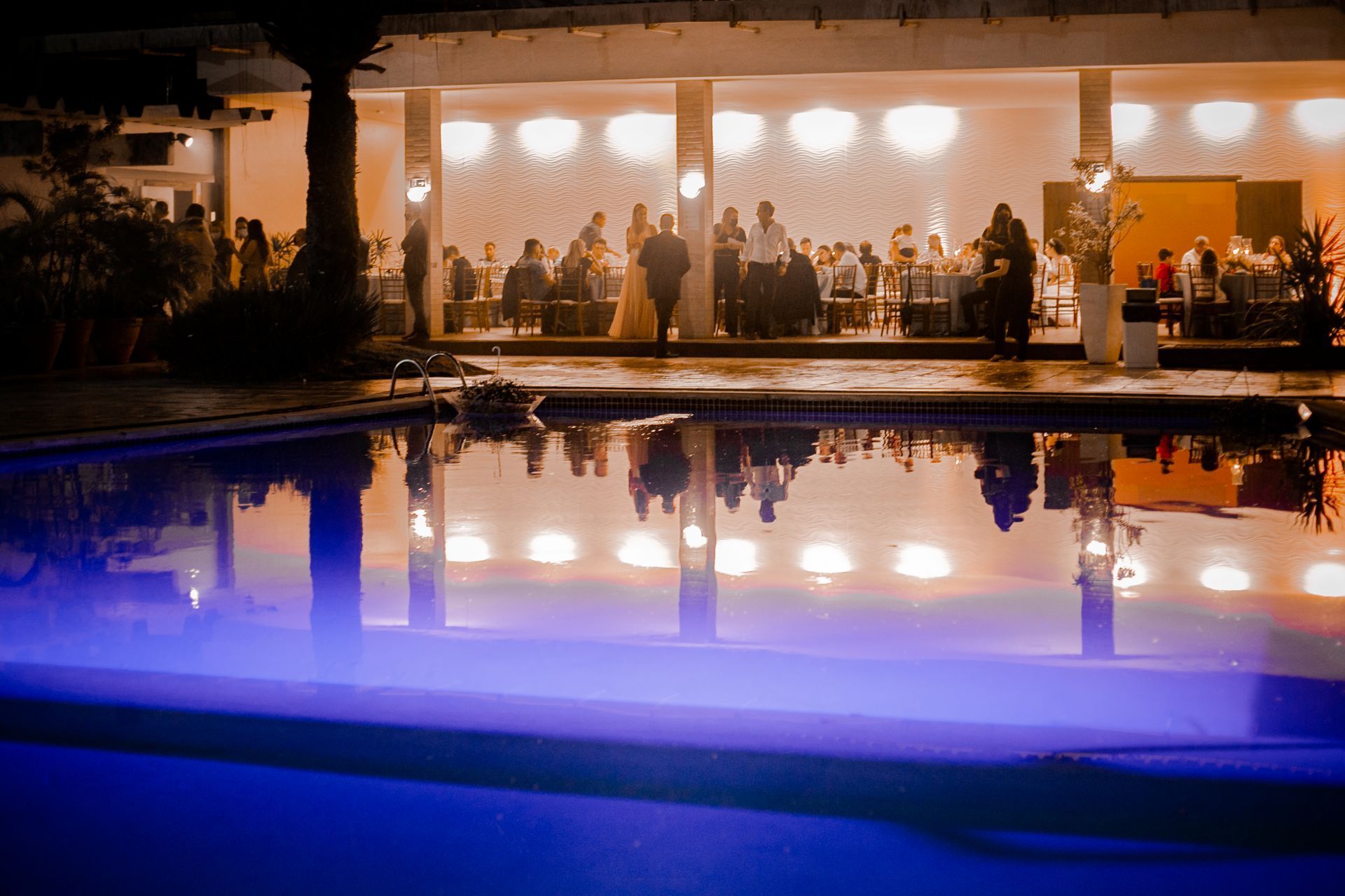 Evento noturno junto à piscina: os convidados, sob uma cobertura iluminada, refletem-se na água.
