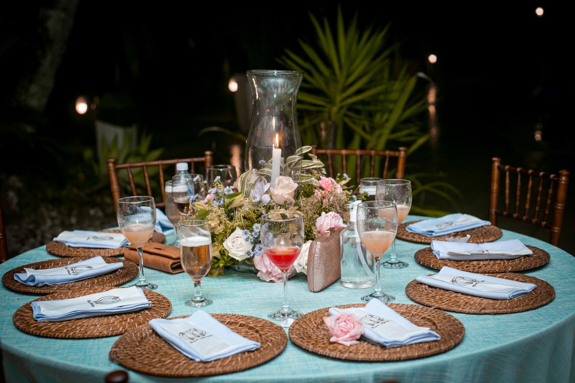 Mesa elegantemente posta com arranjo floral central, toalha de mesa azul-petróleo, jogos americanos e iluminação externa.
