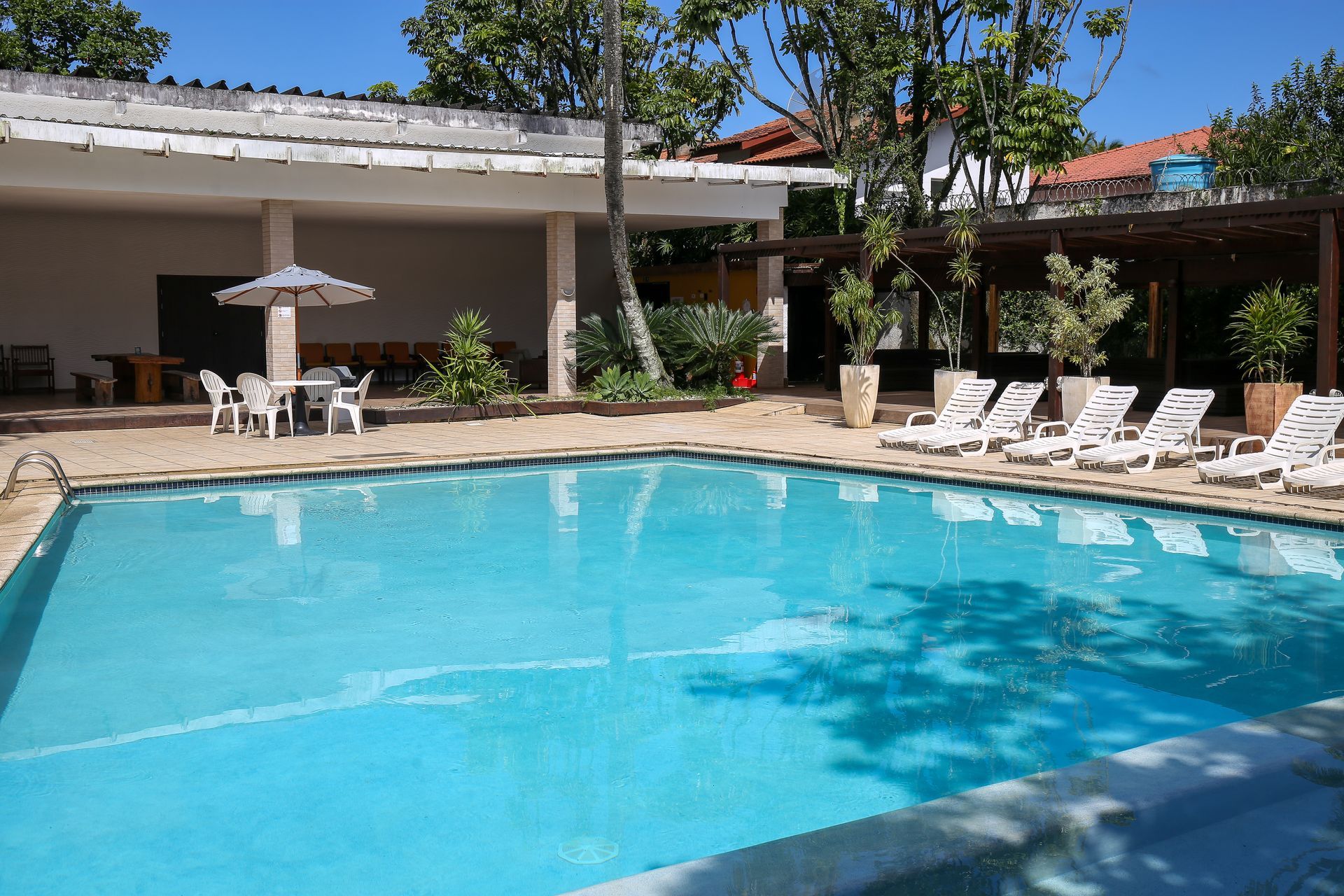 Área da piscina com piscina azul, espreguiçadeiras, pátio e céu ensolarado.