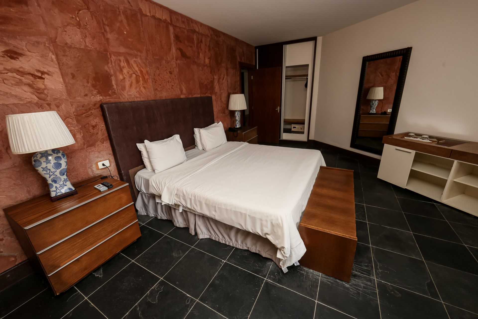 Quarto com cama, mesas de cabeceira e piso de azulejo escuro contra uma parede de pedra vermelha.