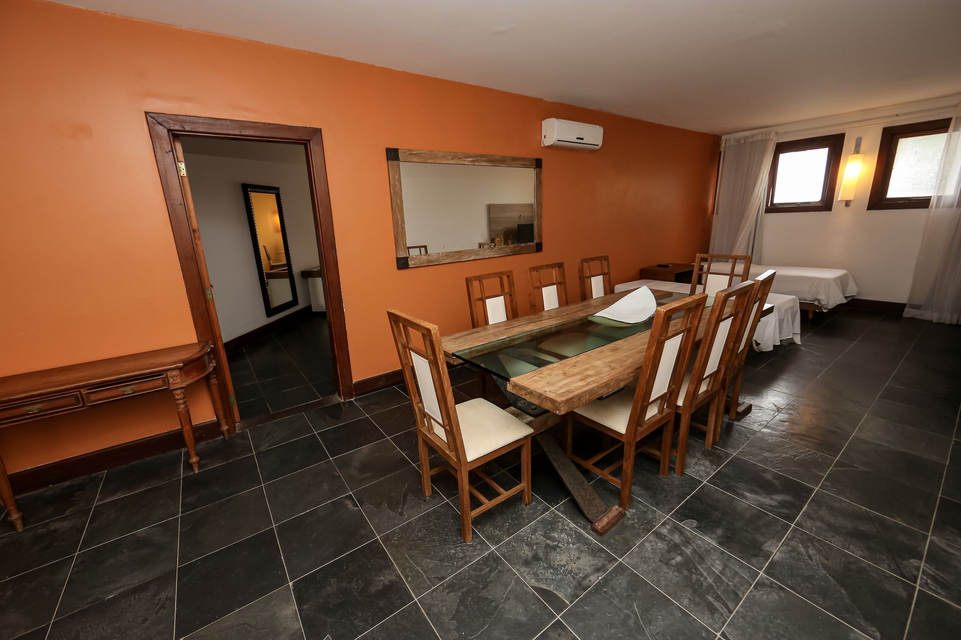 Sala de jantar com mesa comprida de madeira, cadeiras, parede laranja, espelho grande e piso de azulejos pretos.