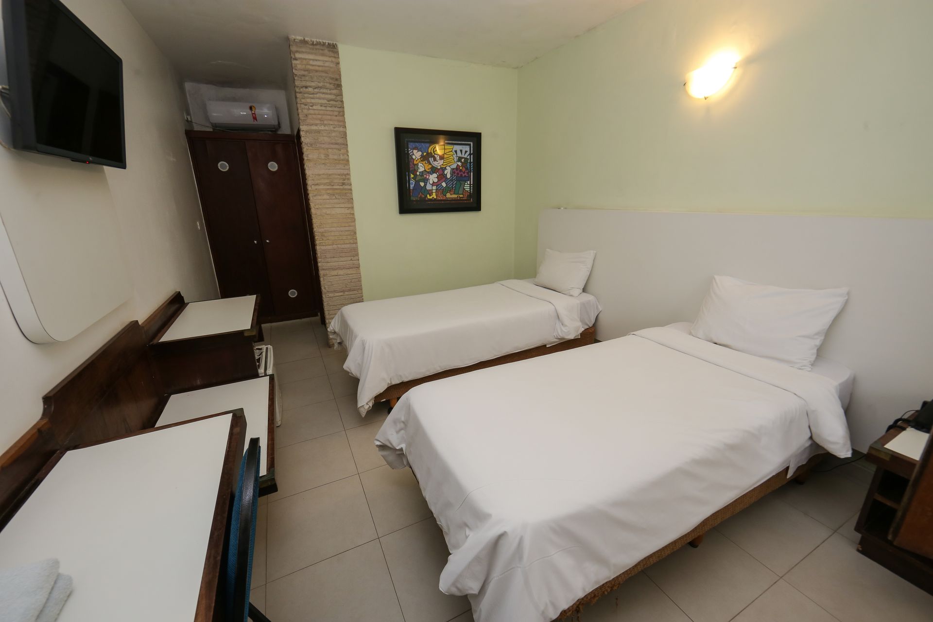 Duas camas em um quarto de hotel com lençóis brancos. Paredes verde-claras, móveis de madeira e um quadro são visíveis.