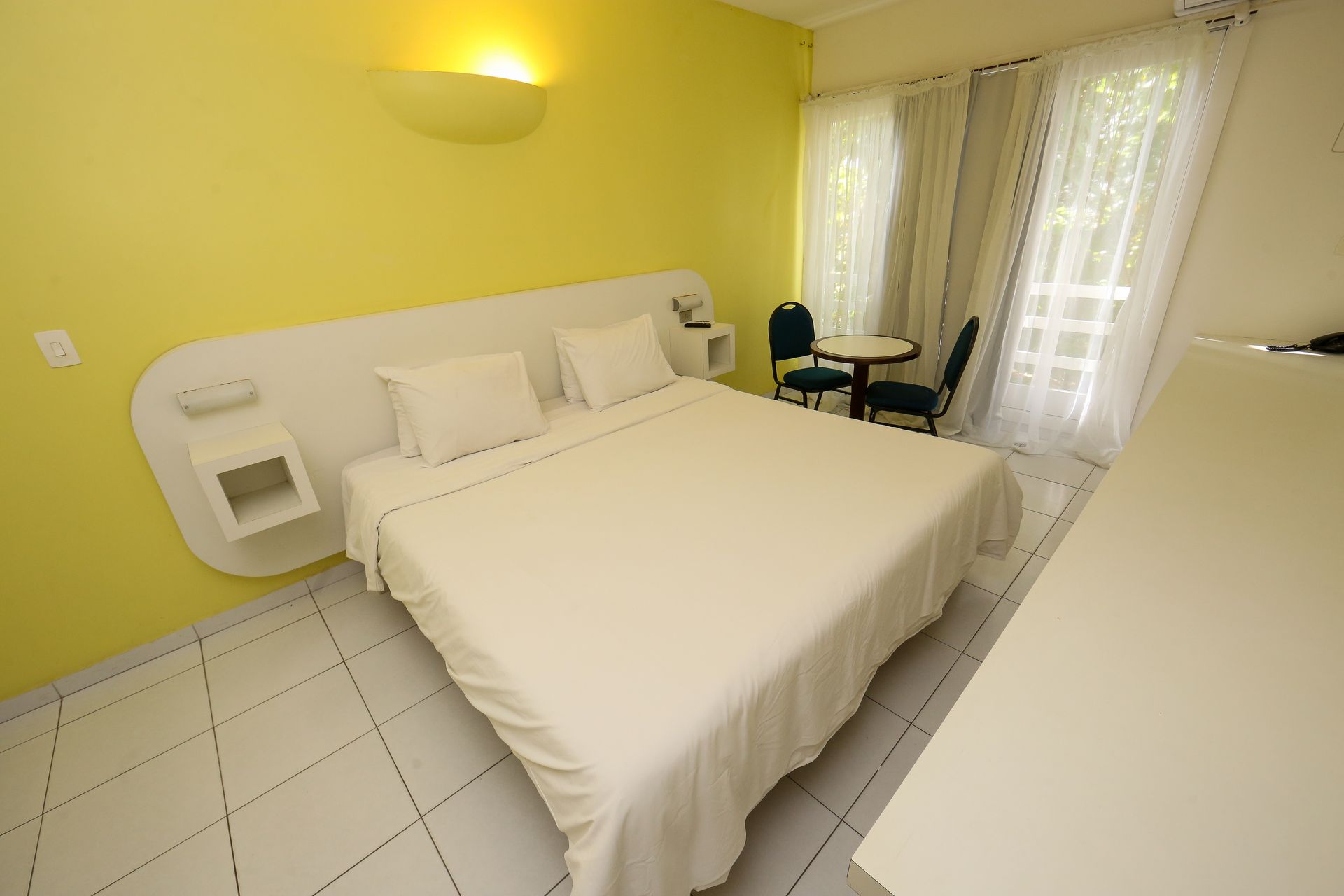 Quarto com cama, mesa e cadeiras. Parede amarela, roupa de cama branca e piso de azulejo.