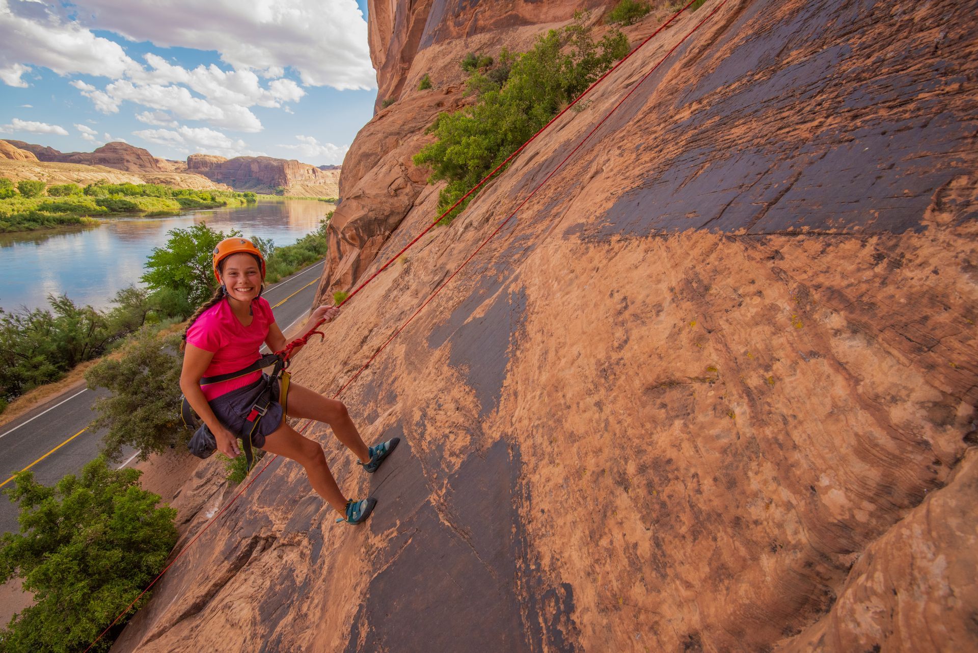 Woman rappelling down a red rock cliff face near a river. Smiling, wearing helmet and harness.