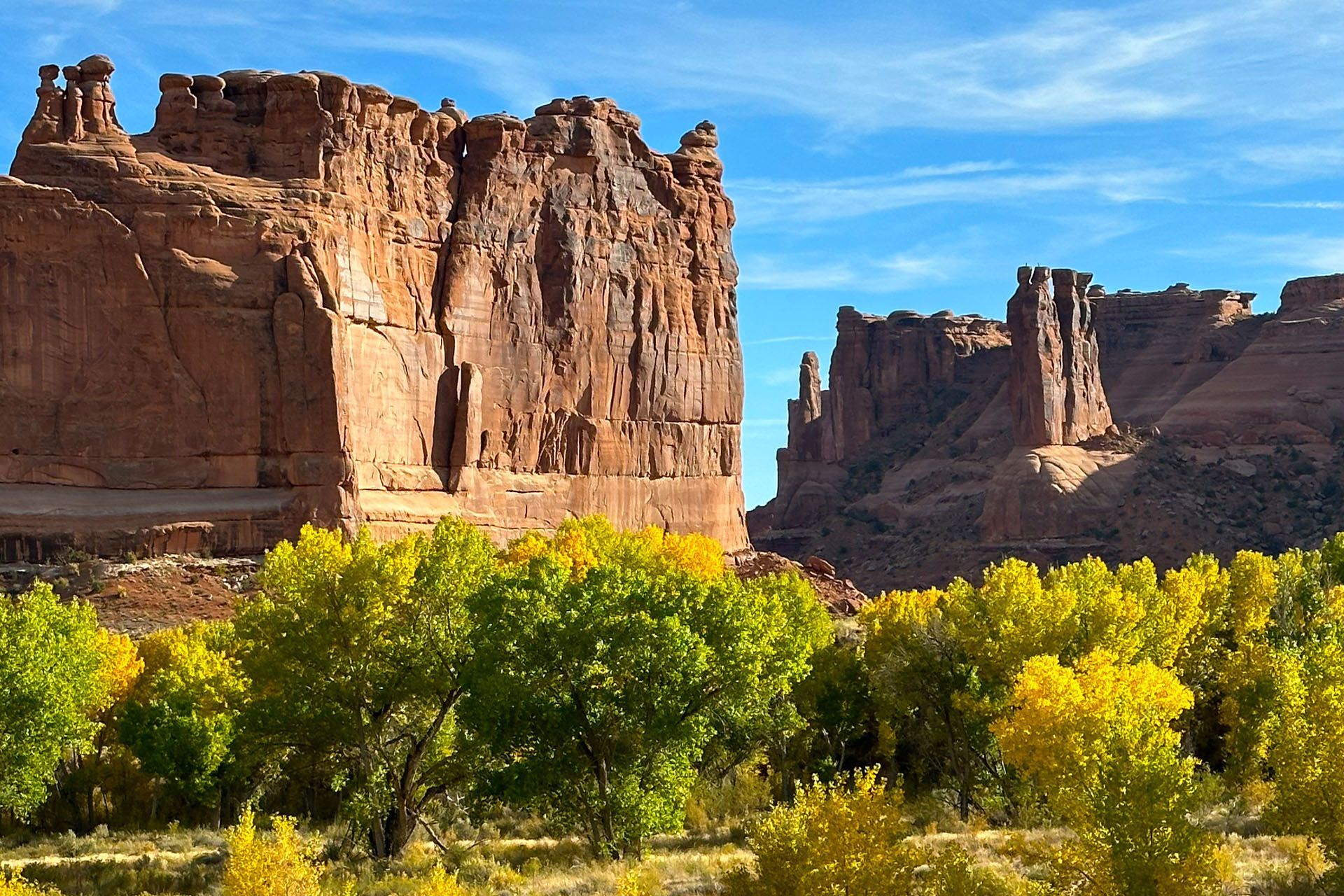 Red rock formations tower over a line of trees with green and yellow leaves against a blue sky.