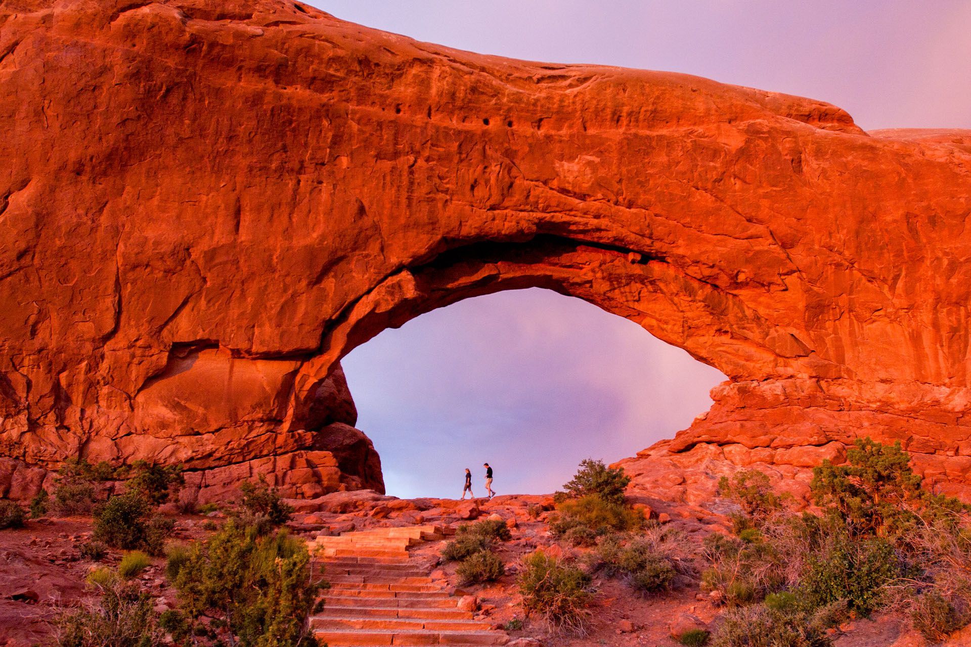Two people walk below Window Arch in Arches National Park as the sunset reflects off the rock and sky.