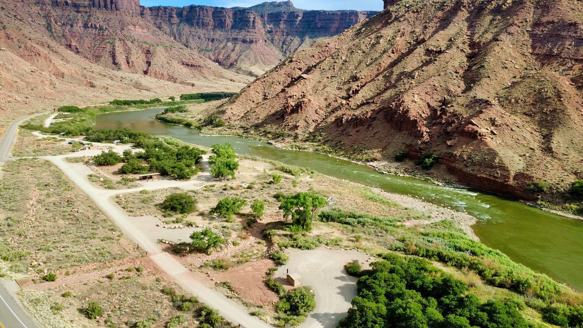 River flowing through a canyon; campground along the bank with trees, red rock cliffs in background.
