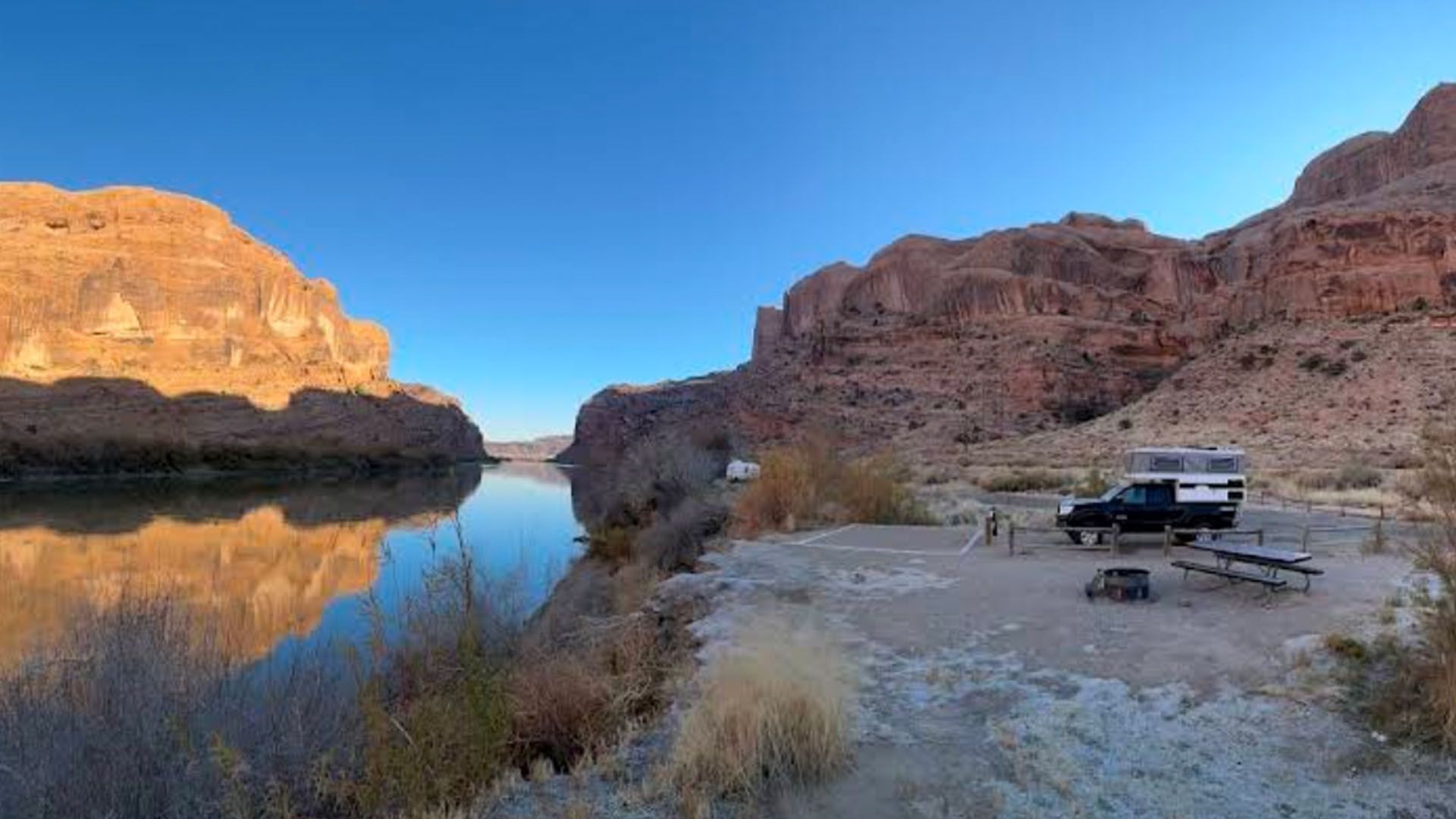A camper truck parked by a river with rocky cliffs, bright blue sky, and calm water reflecting the surroundings.