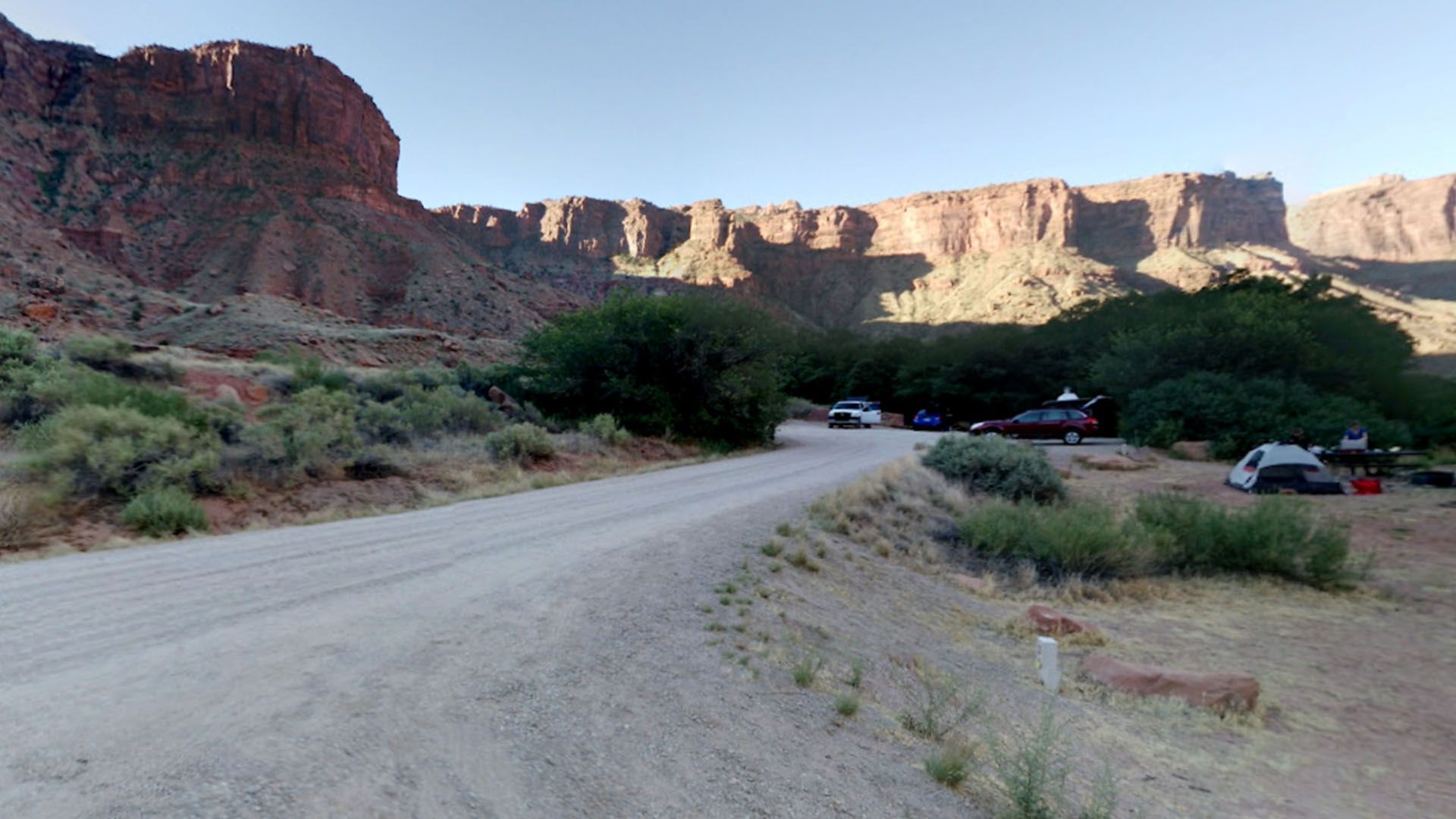 Dirt road leading to a campsite near a red rock cliff face, with parked cars and tents visible.