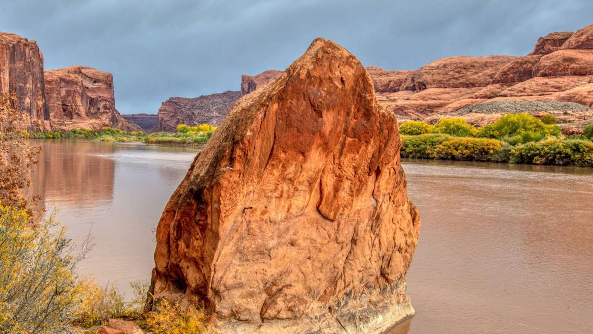 Red rock formation in front of a river, with canyon walls and cloudy sky in the background.