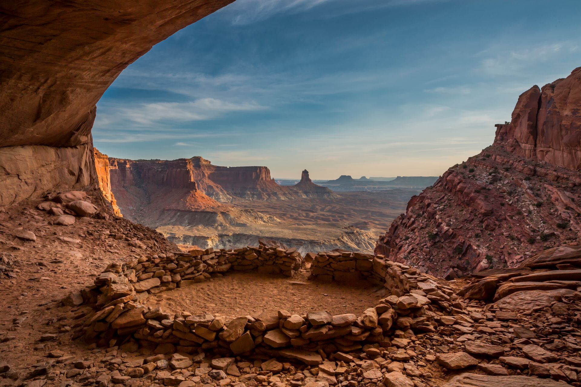 Stone structure under a rock overhang overlooking a vast desert landscape, red rock cliffs, blue sky.