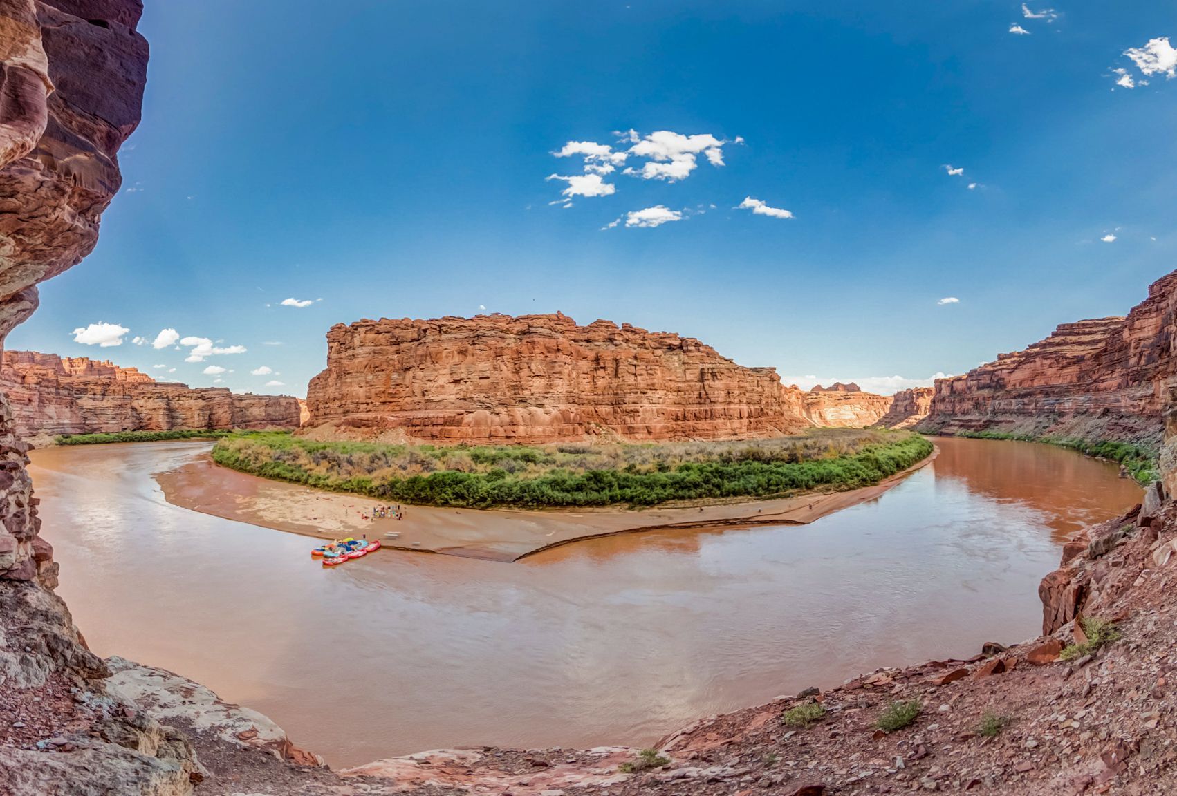 River flows around a red rock island under a blue sky. Raft visible at a beach camp.