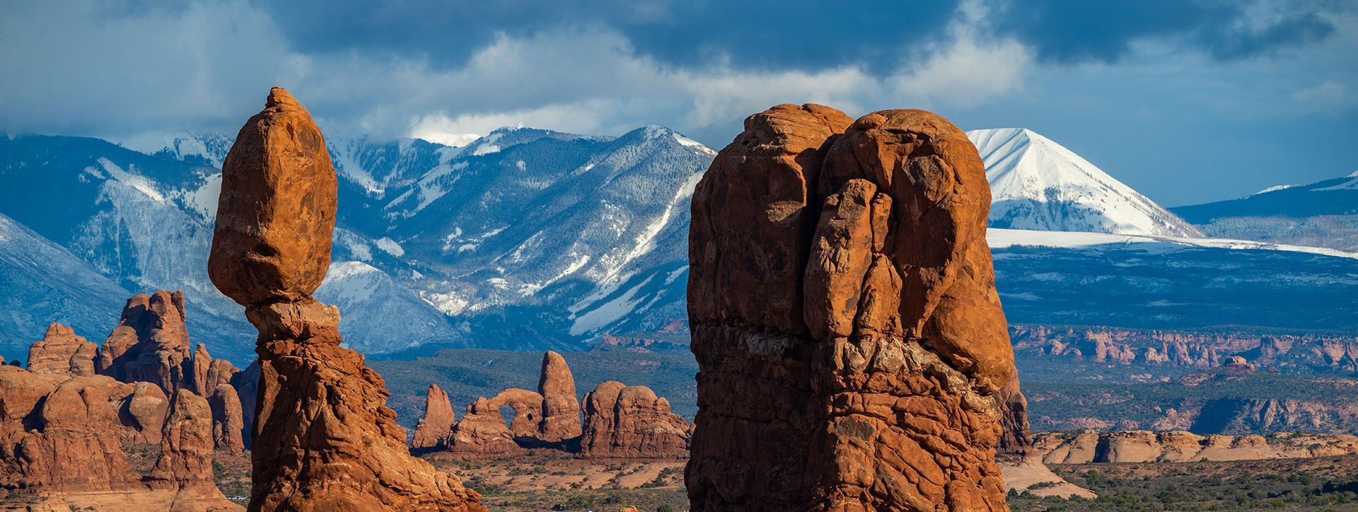 Balanced Rock and other spires tower in the foreground with snow-covered mountains and clouds in the distance