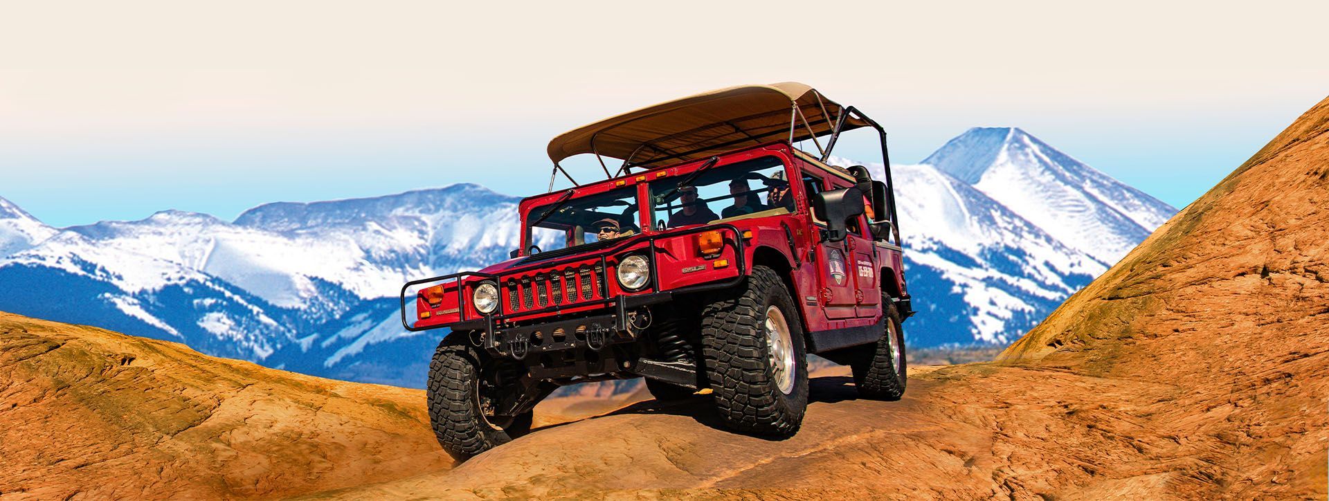 Red Hummer drives up a sandstone hill in a mountainous setting with a snow-capped mountain backdrop.