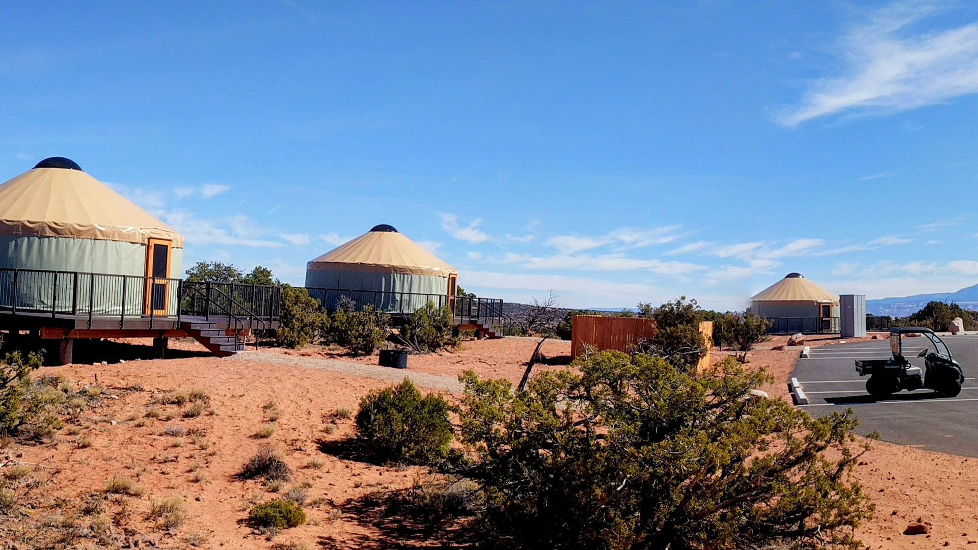 Three yurts in a desert landscape under a blue sky. A golf cart is parked nearby.