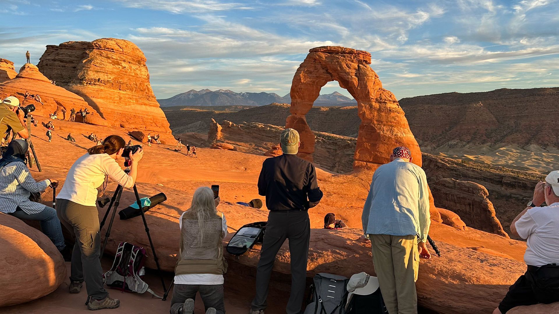 People at Delicate Arch, Utah, photographing the orange sandstone formation at sunset.