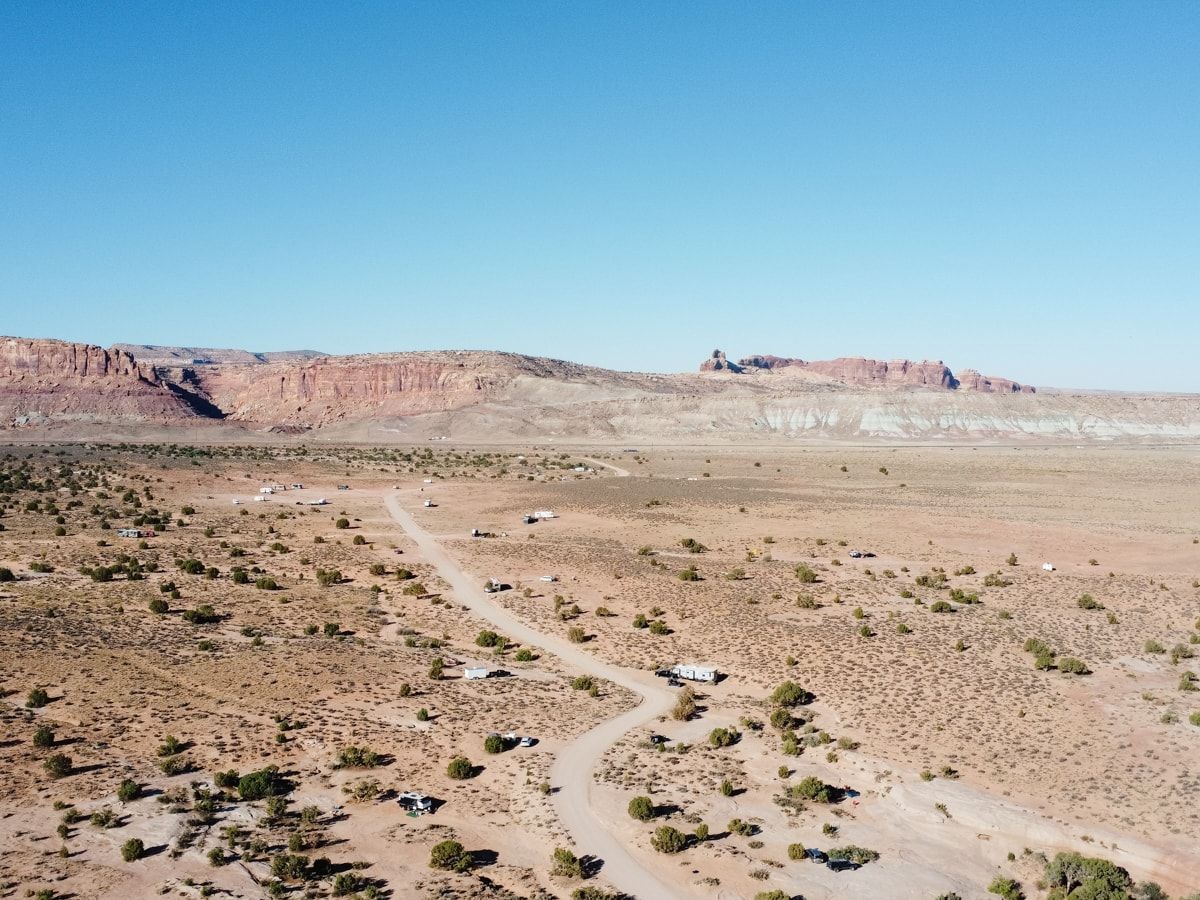Dirt road through arid landscape; red rock formations and blue sky in background.