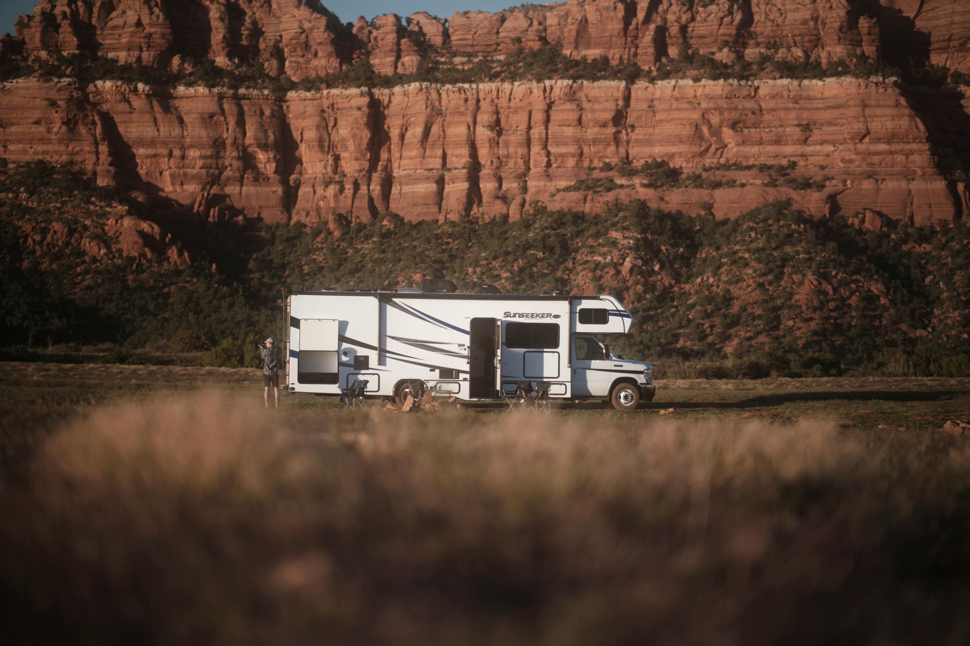 White RV parked in front of a red rock cliff, in a field with dry grass.