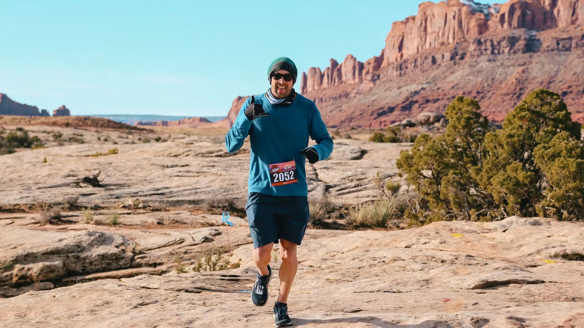 Runner on a desert trail, wearing blue, giving a thumbs up, with red rock formations in the background.