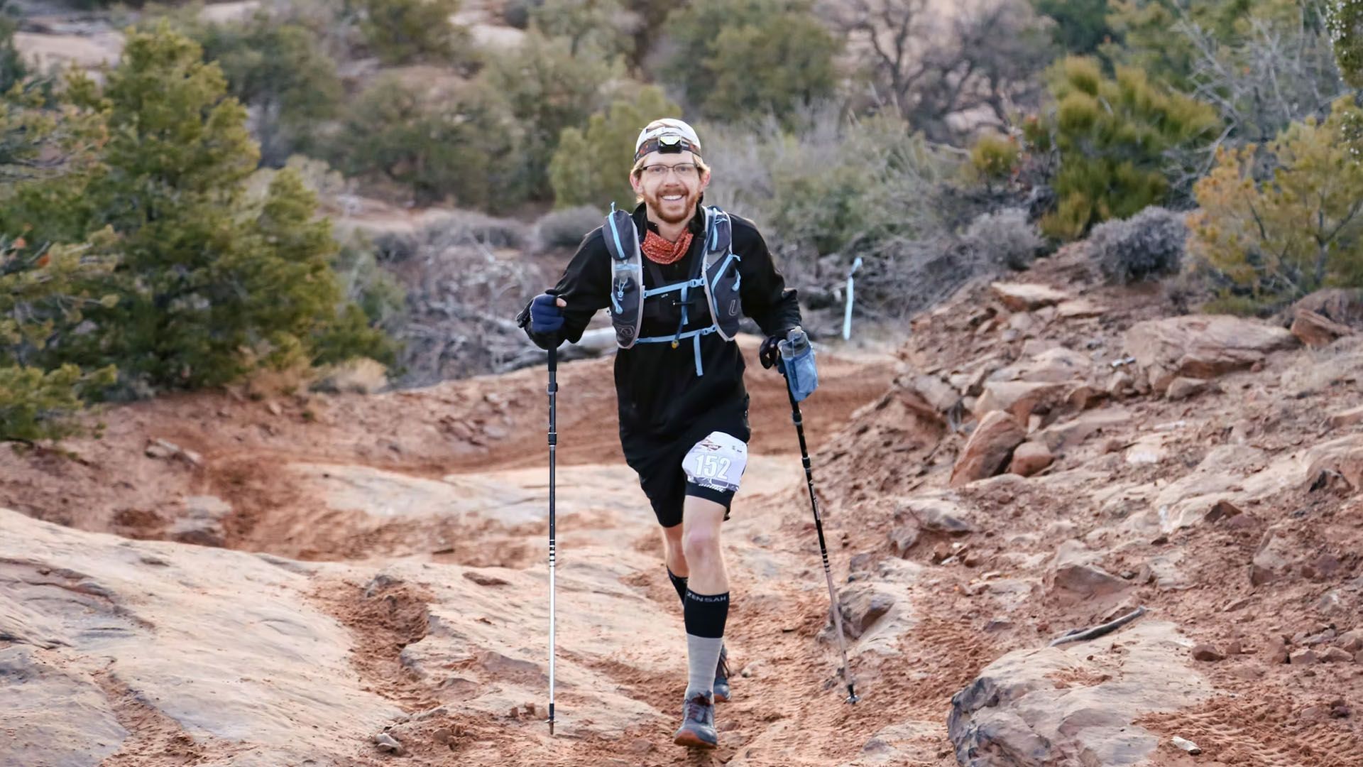 Man trail running uphill on a rocky, reddish path, using trekking poles. He wears black clothing.