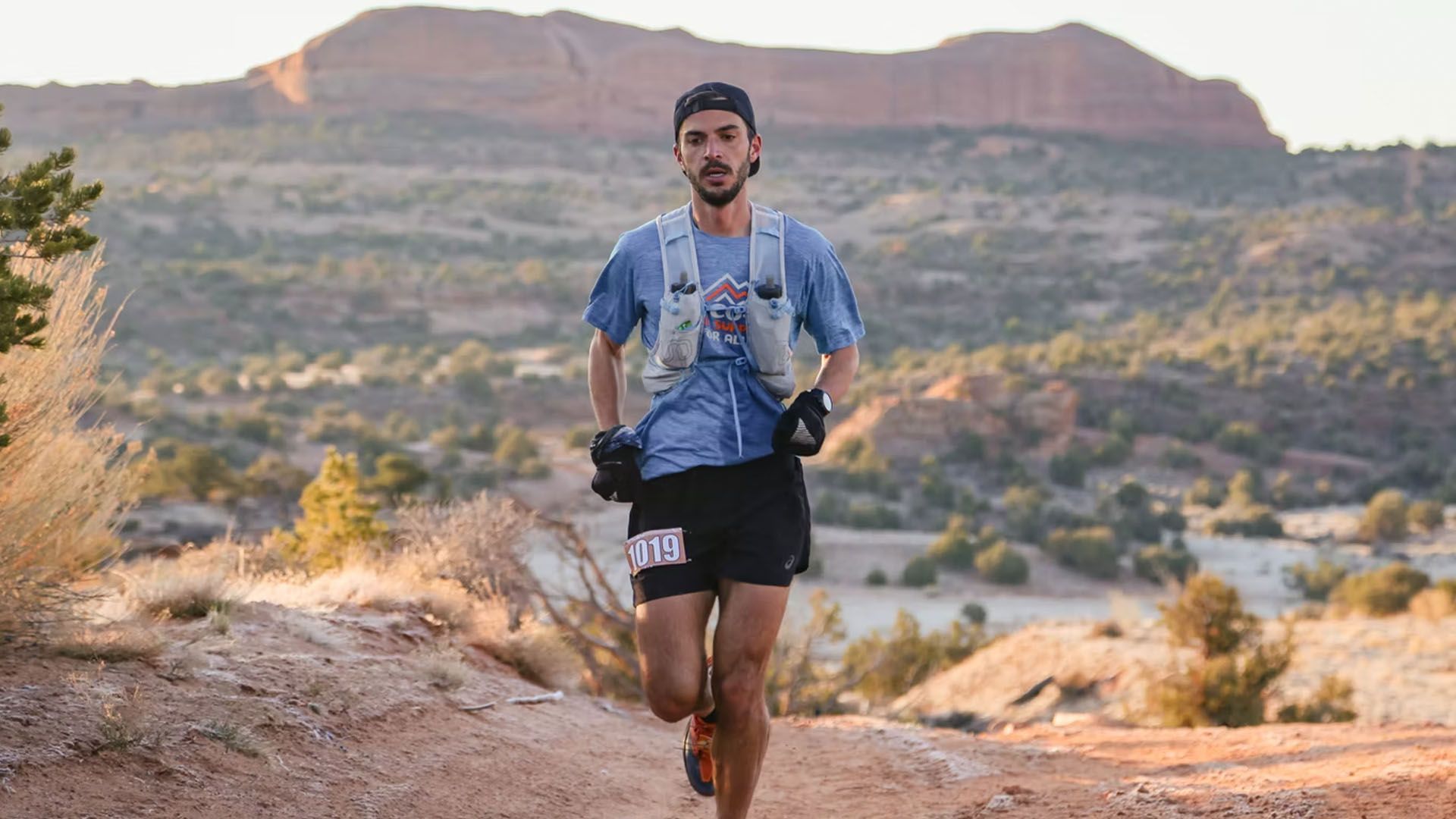 Man trail running on dirt path with mountain backdrop. He wears a hydration pack and shorts.