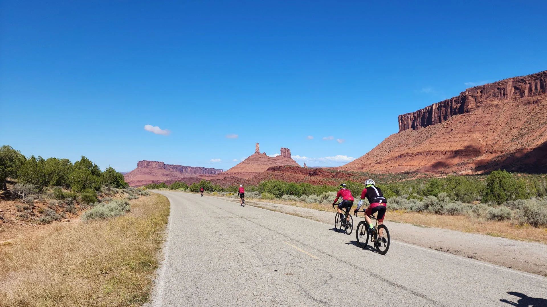 Cyclists ride along a dirt road with red rock formations and blue sky in the background.
