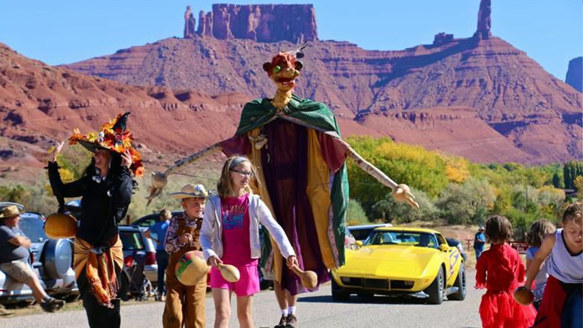 Parade with giant puppet, costumes, classic yellow car, red rock formations in background.