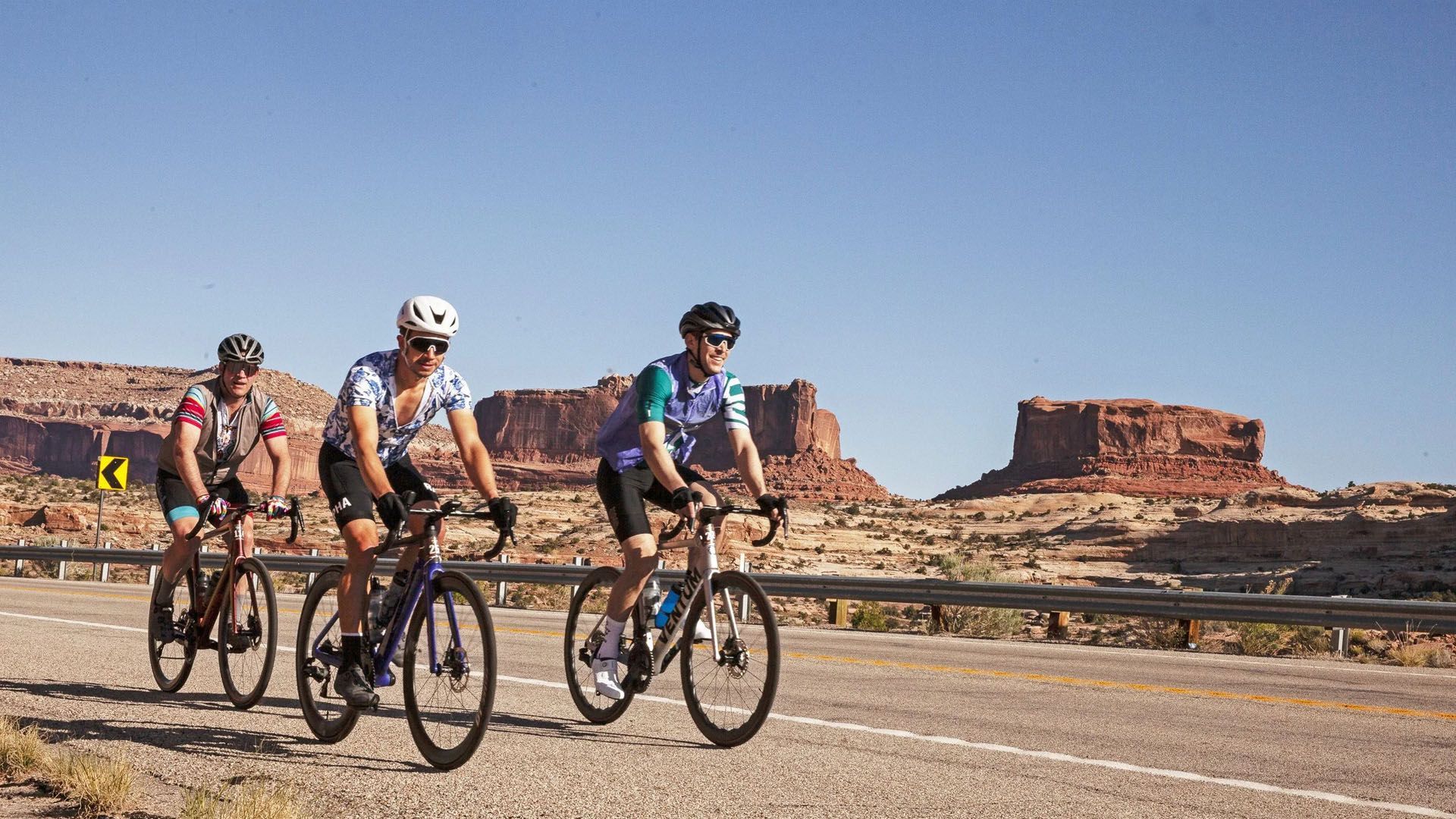 Three cyclists ride road bikes on a desert highway with red rock formations under a blue sky.