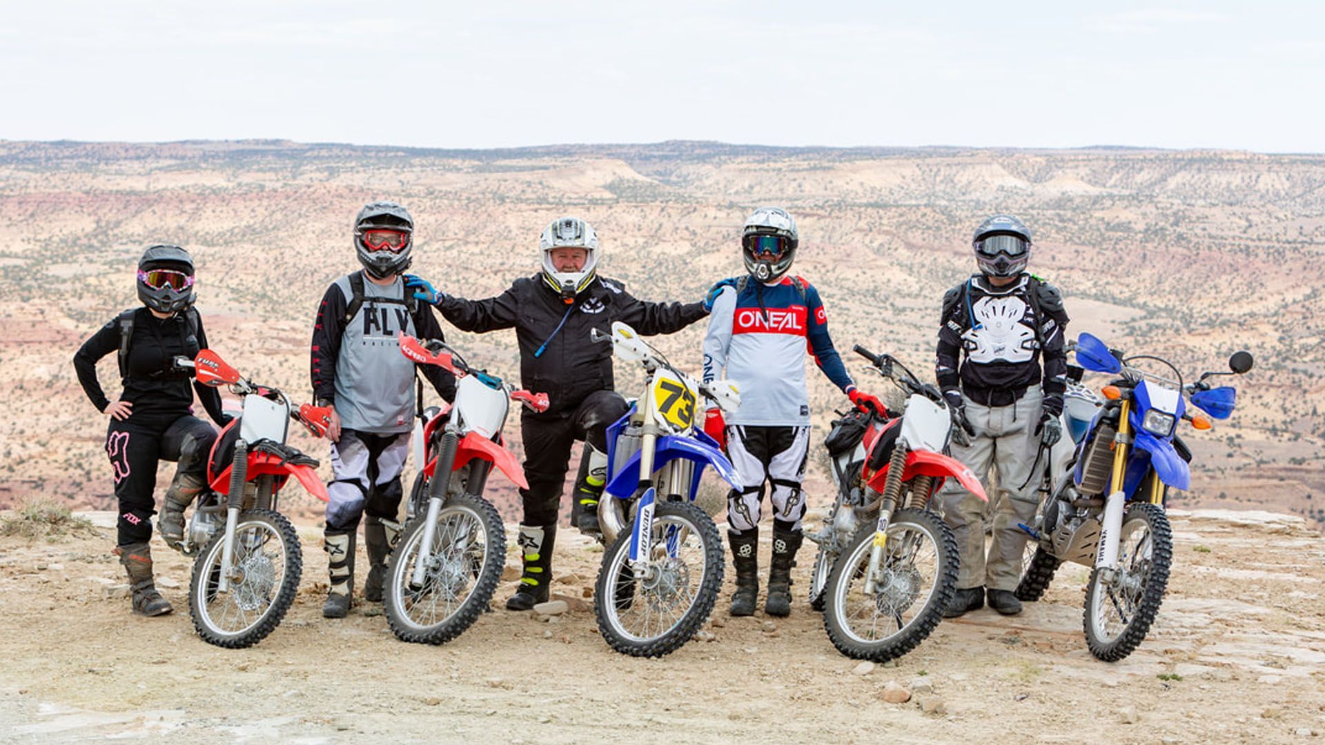 Group of dirt bike riders pose with their bikes in a desert landscape.