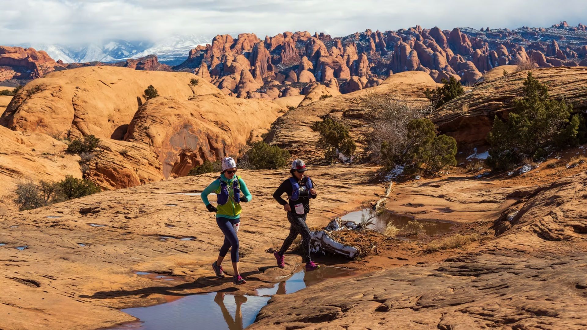 Two trail runners in sportswear on a red rock desert trail. Mountains in the background, puddles in the foreground.