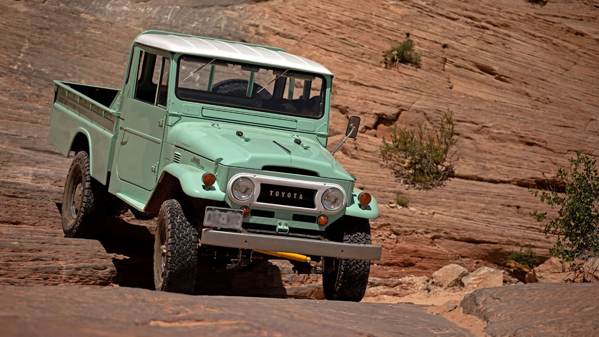 Vintage mint green Toyota Land Cruiser pickup truck off-roading on a rocky, desert path.