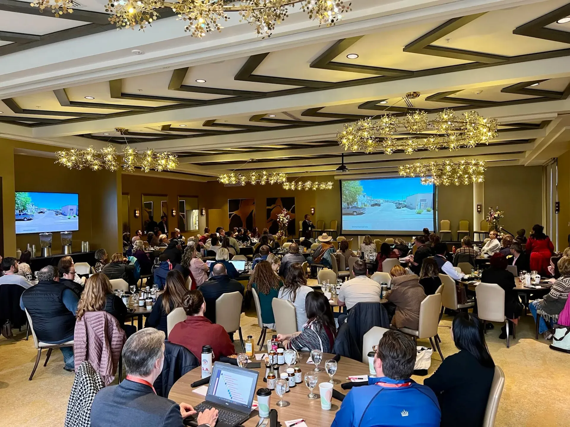 Conference attendees seated at tables, watching presentations on screens in a large, ornate ballroom.