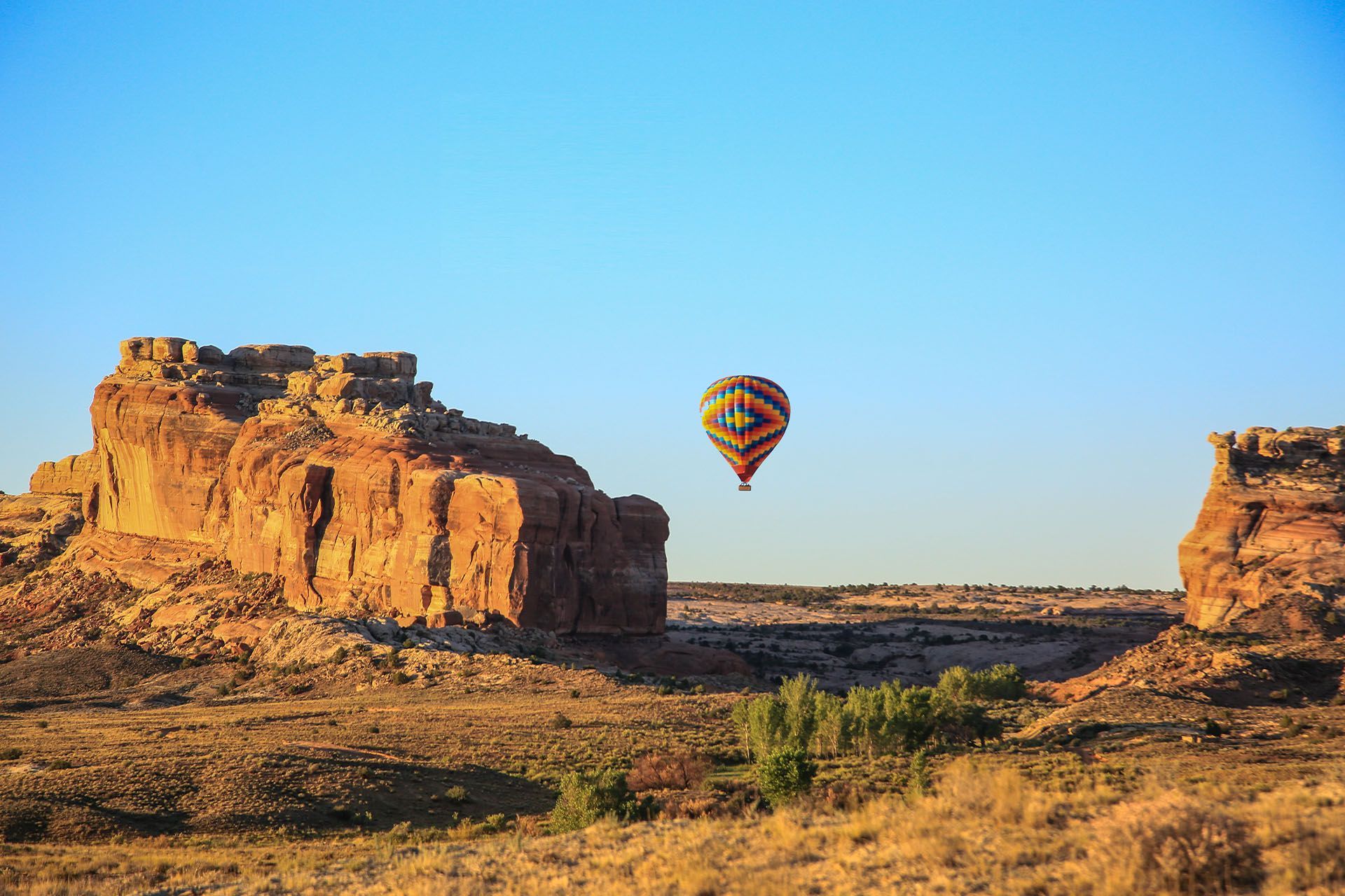 A hot air balloon floats over the Moab desert with tall plateaus in the distance.