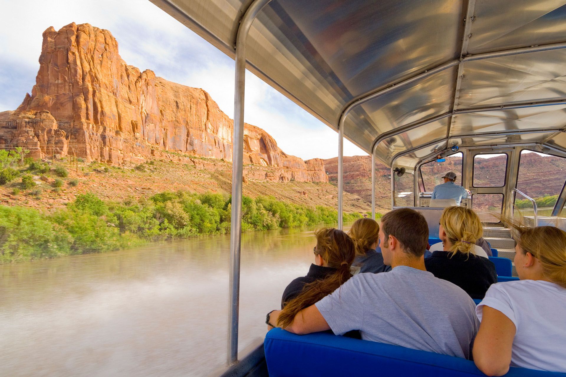 Jet boat on river with passengers, red rock formations in background.