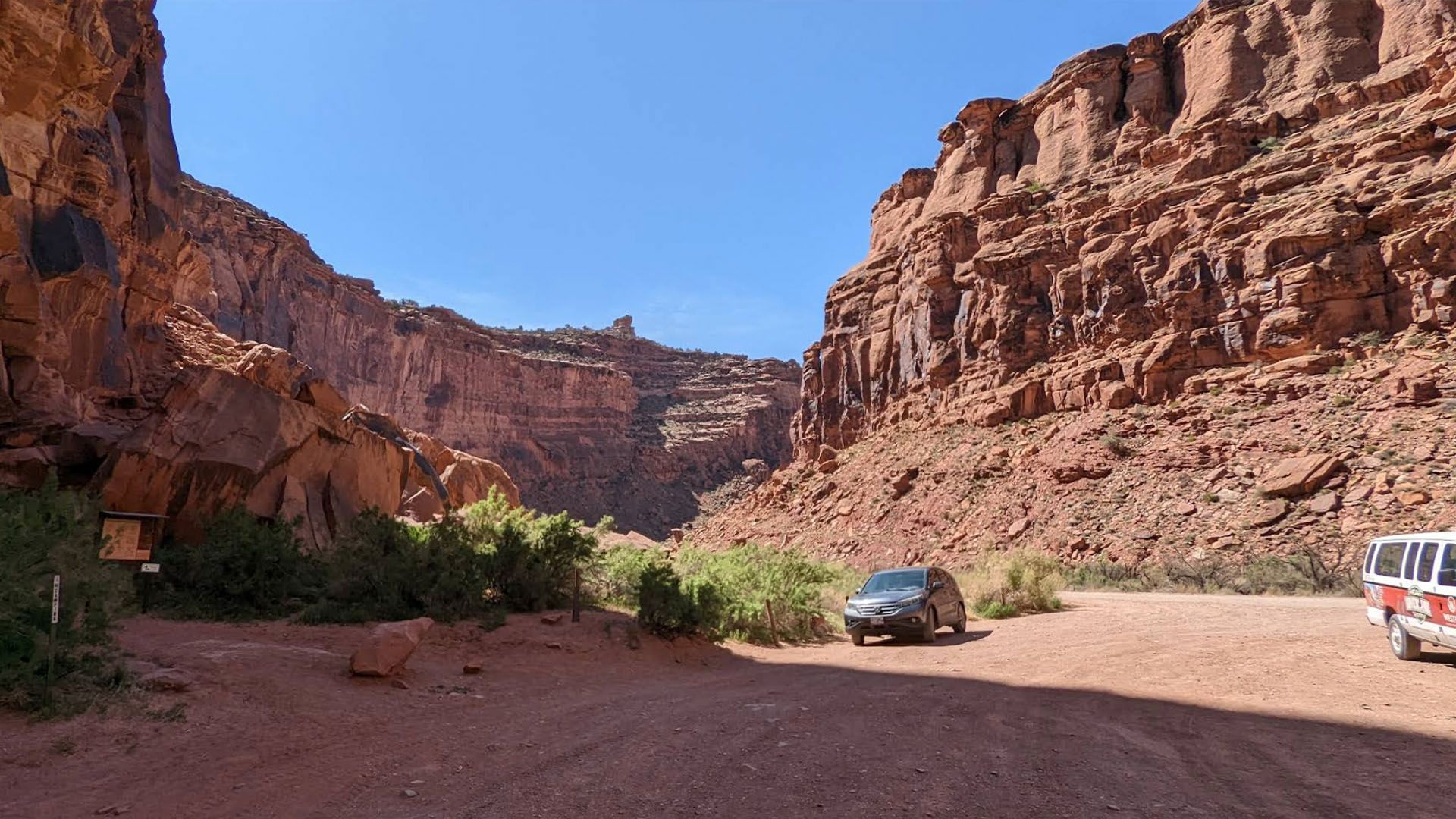 Red canyon with a vehicle parked on a dirt road; clear blue sky.