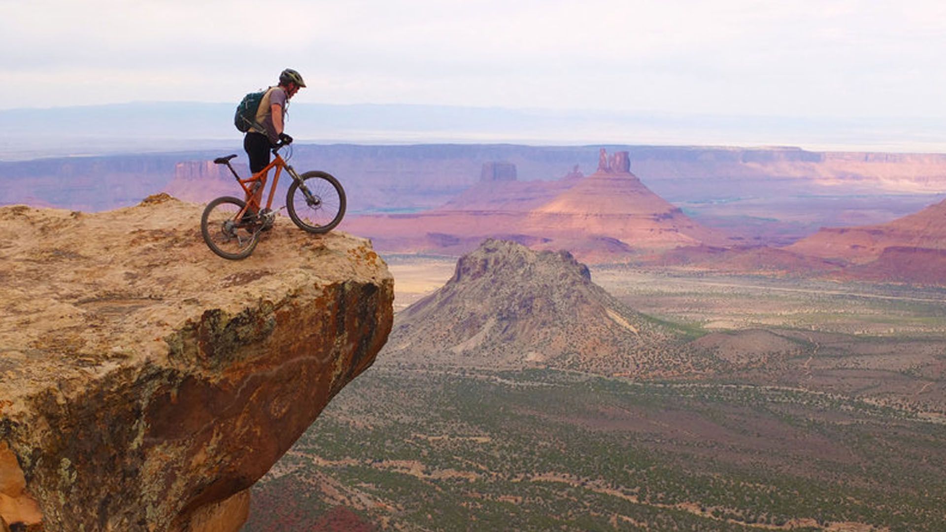 Man on mountain bike at cliff edge, overlooking red rock canyon landscape.