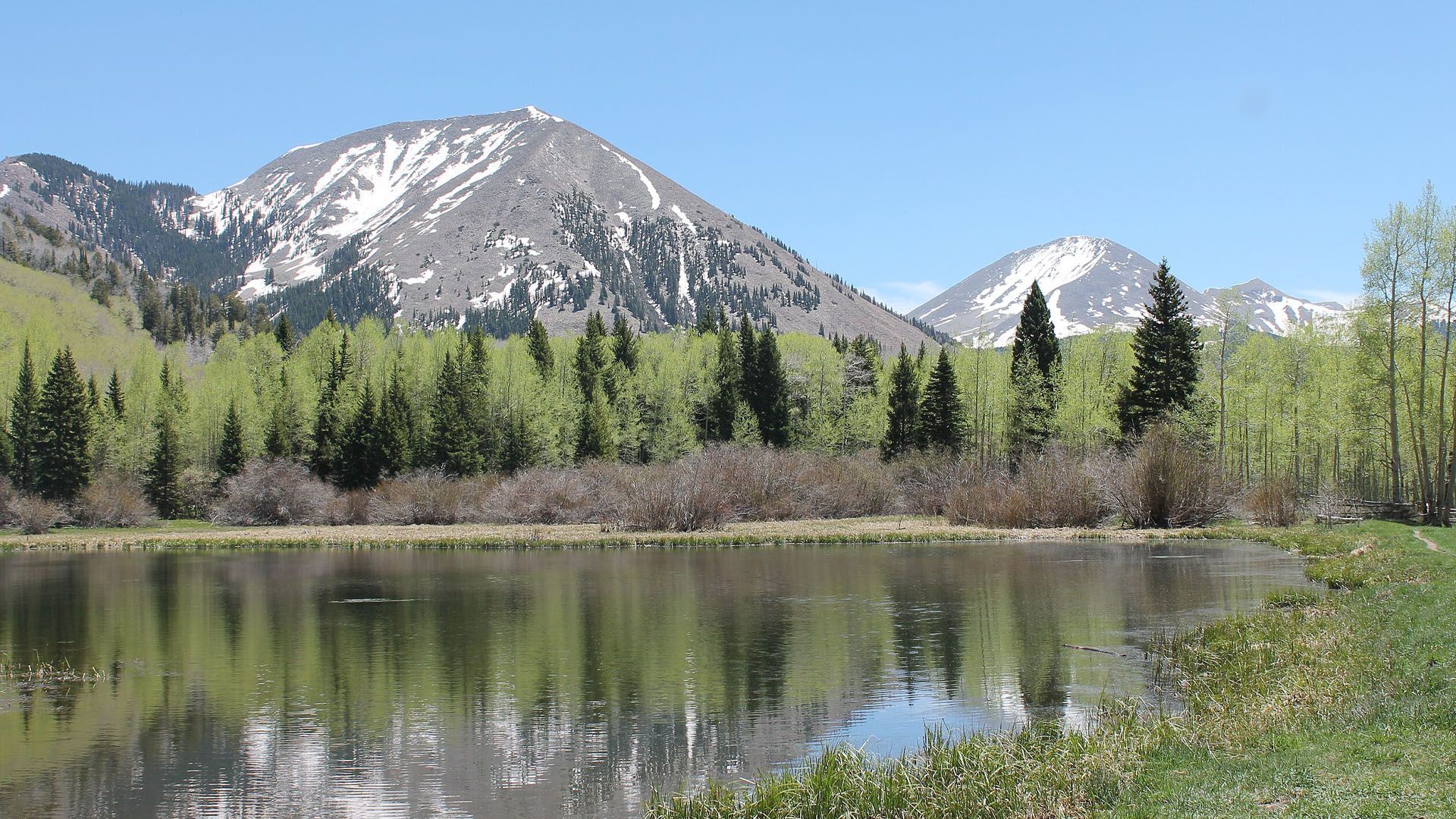 Lake reflecting mountains with patches of snow, surrounded by green trees.