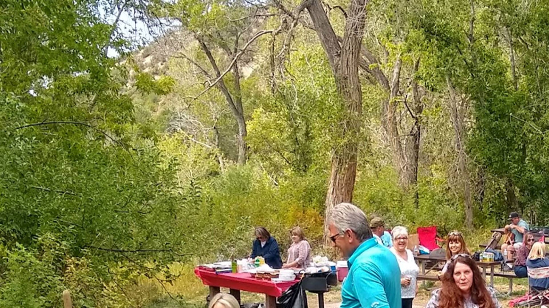 People gathered for a picnic in a park with a red table of food surrounded by trees.