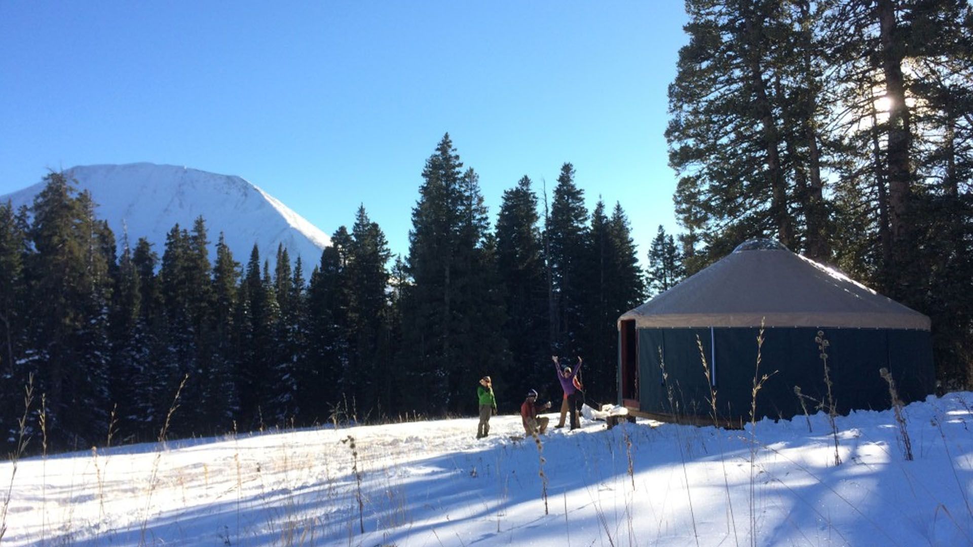 Snowy scene with yurt, people near a fire, and mountain in background.