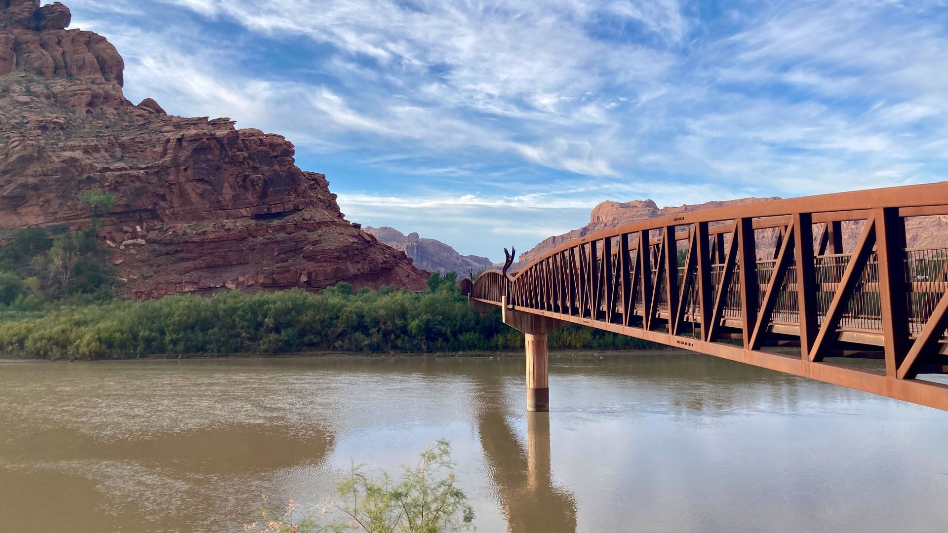Bridge over river with red rock cliffs in the background under a blue sky.