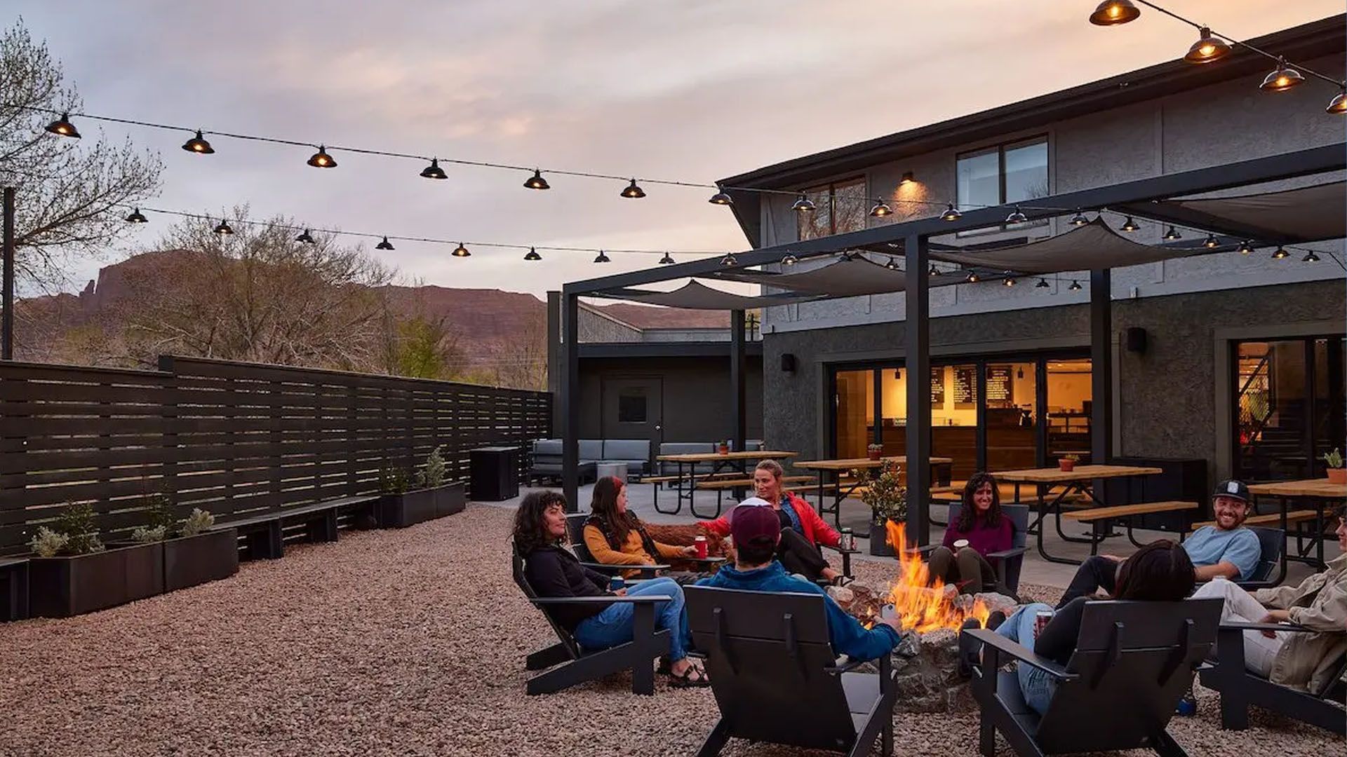People gathered around a fire pit on a patio, under string lights. Building and mountains in the background.